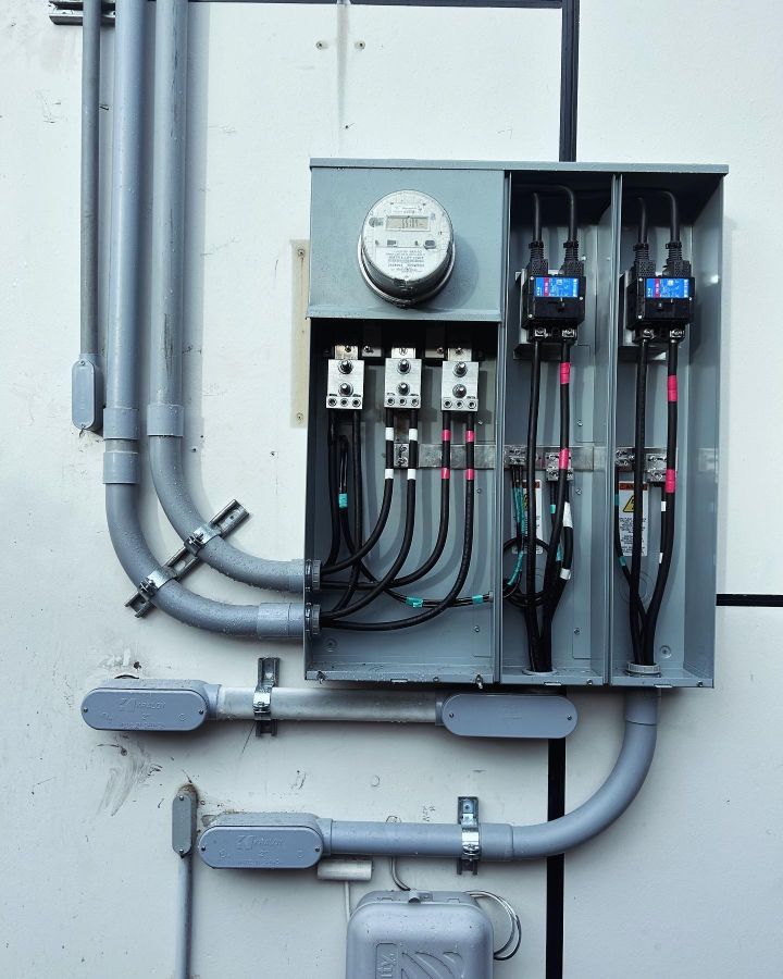 Electrical meter and breaker panel with conduit and wiring on a gray wall.