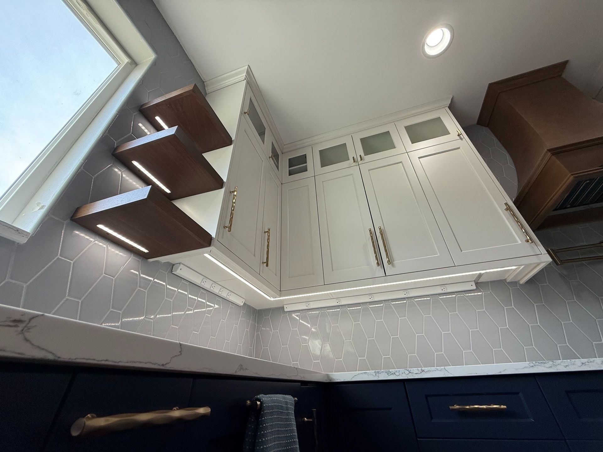 Tilted view of a modern kitchen with white cabinets, dark blue drawers, a window, and floating shelves.