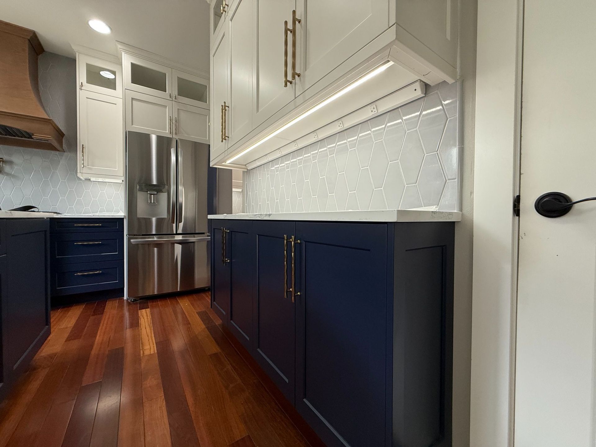 Modern kitchen with dark blue cabinets, stainless steel fridge, white uppers, and wood floors
