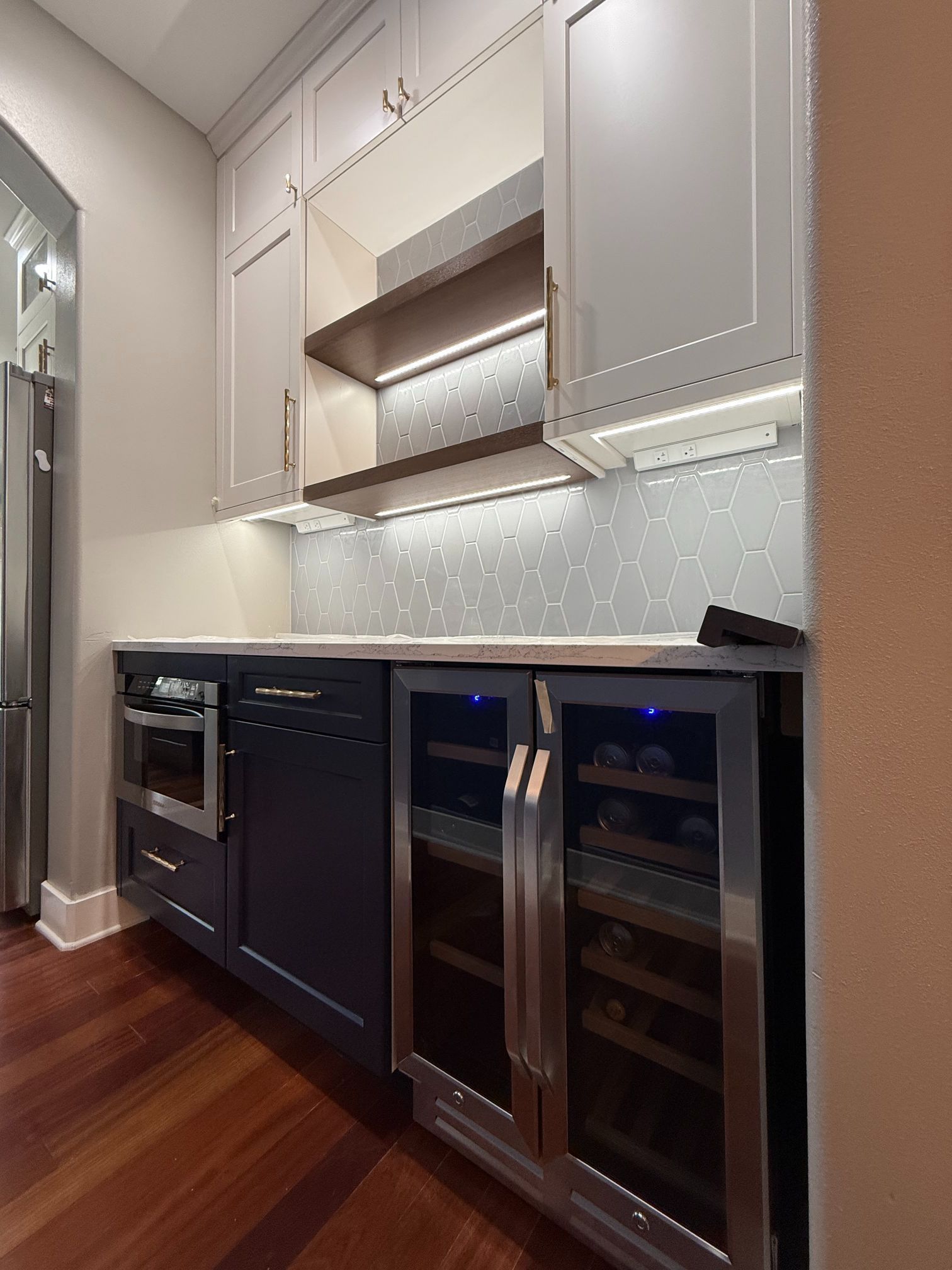 Modern kitchen with gray cabinets, wood floor, under-cabinet lighting, oven, and glass-front wine cooler.