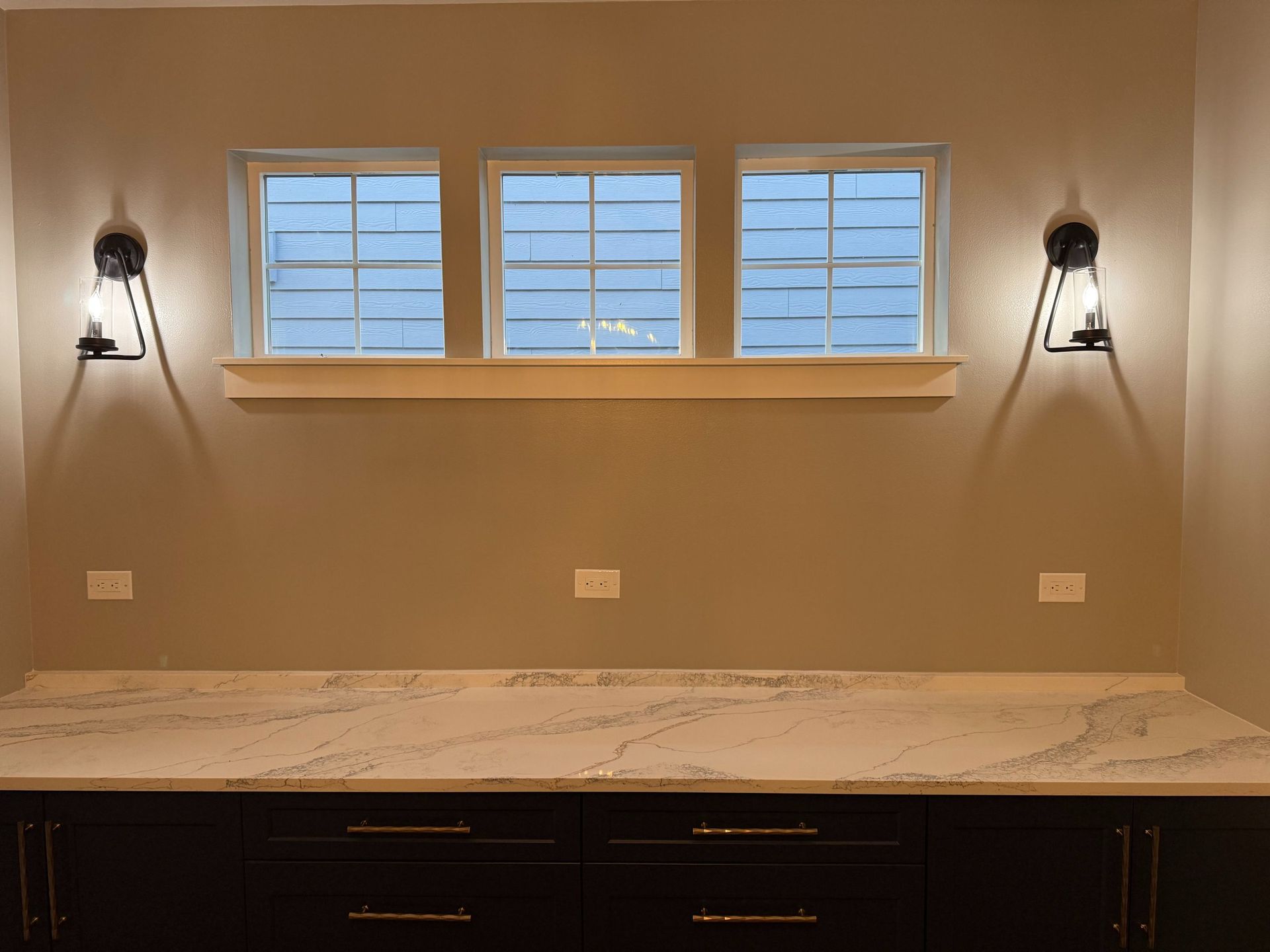 Bathroom vanity with marble countertop, three windows, and wall sconces on either side.