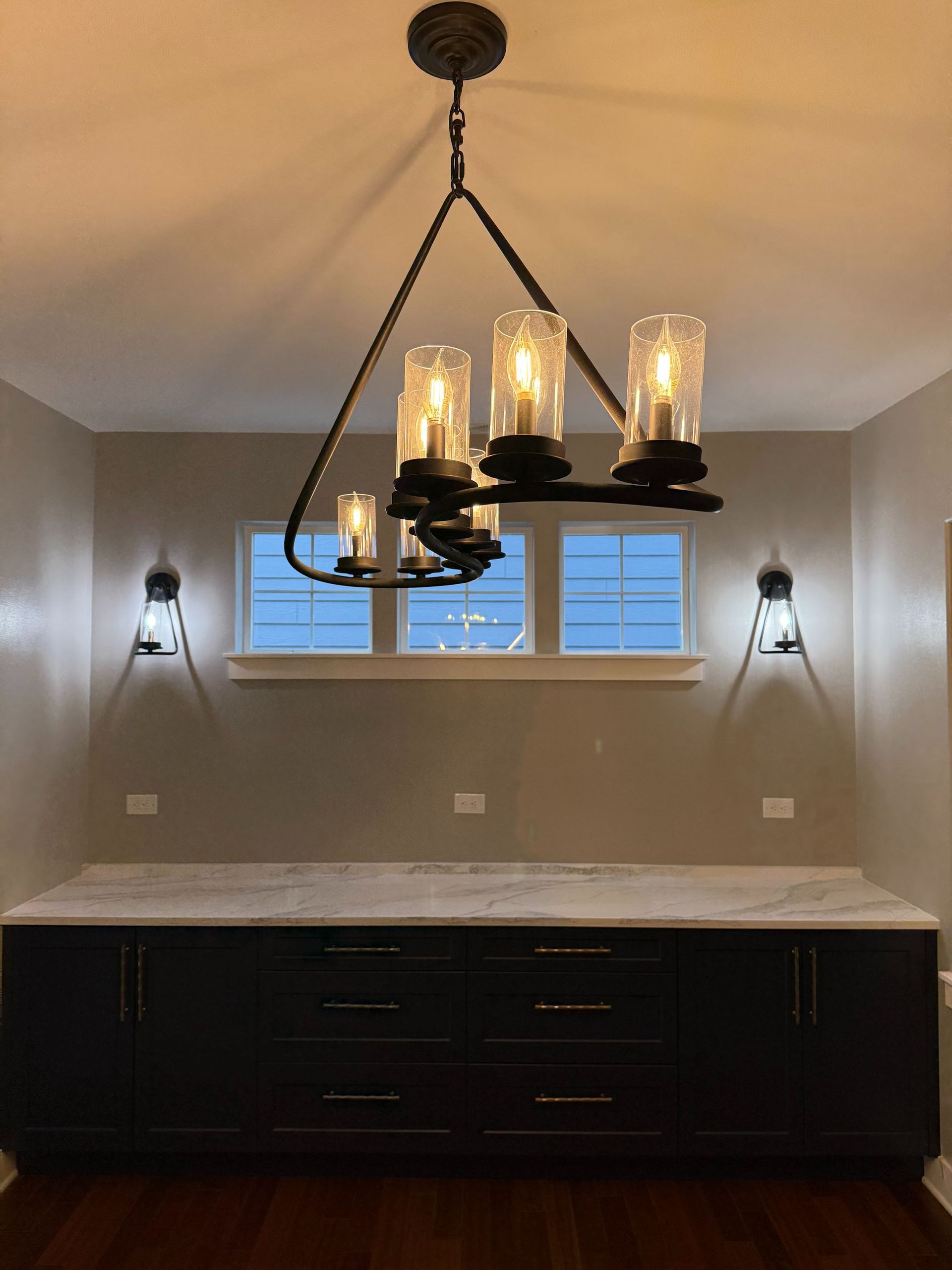Dining room with a dark chandelier, white stone countertop, dark cabinets, and wall sconces over a window.