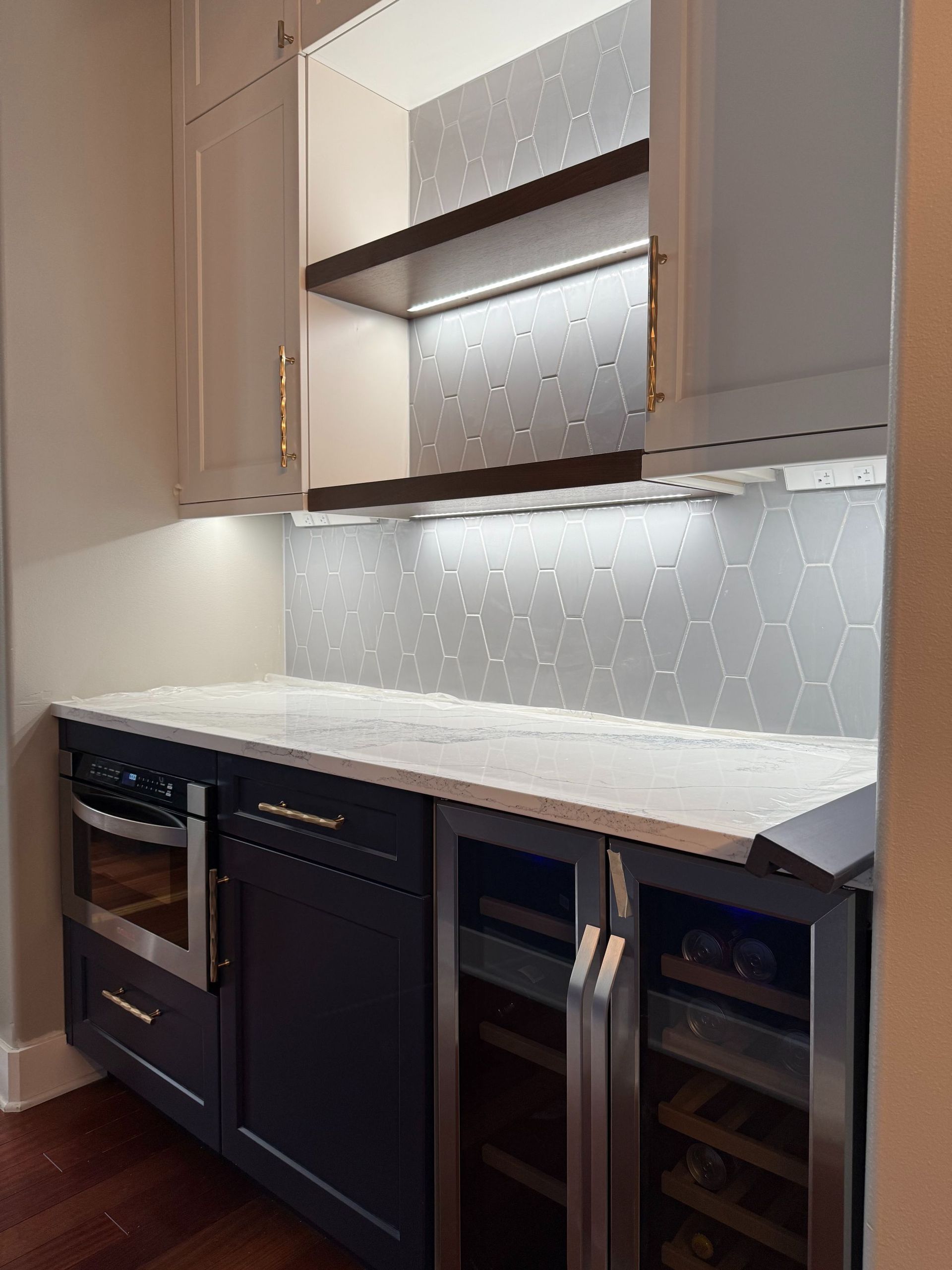 Modern kitchen with beige cabinets, white countertop, under-cabinet lighting, oven, and glass-front wine cooler.