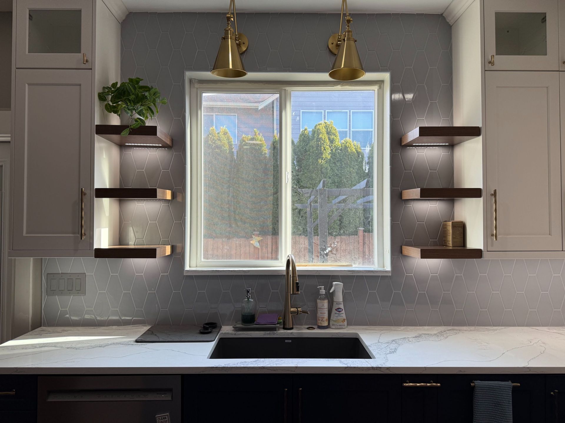 Modern kitchen sink under a window, with floating shelves, gray cabinets, and two brass pendant lights.