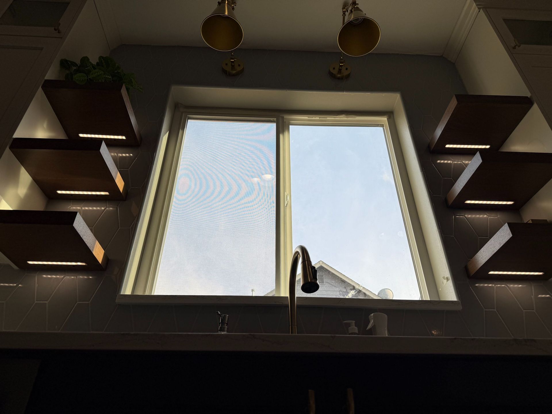 Kitchen sink beneath a window, framed by dark shelves and two hanging pendant lights.