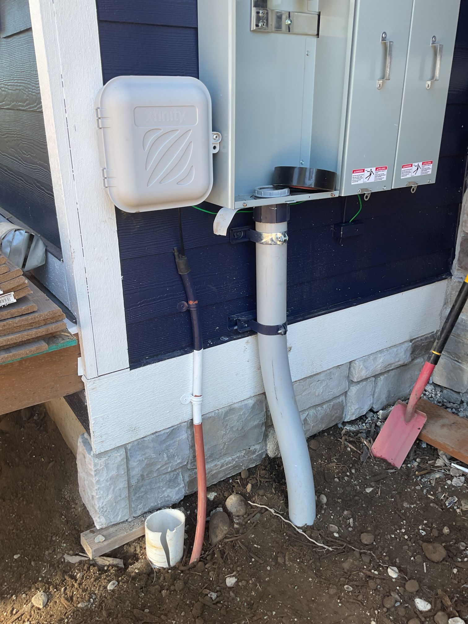 Utility boxes and pipes on a house exterior with a red hose and a pink shovel in dirt below