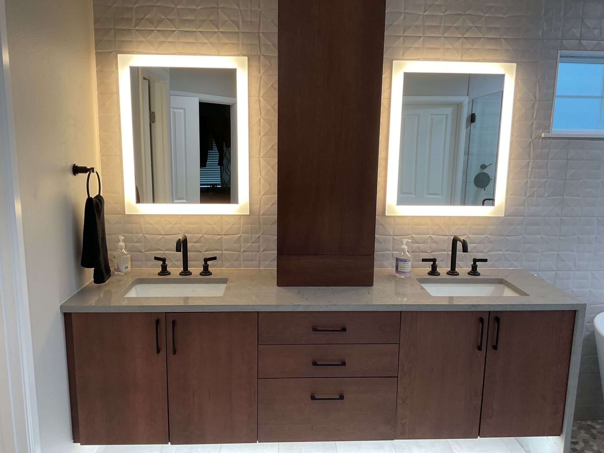 Double vanity bathroom with wood cabinets, marble countertop, and two lit mirrors against white tile walls