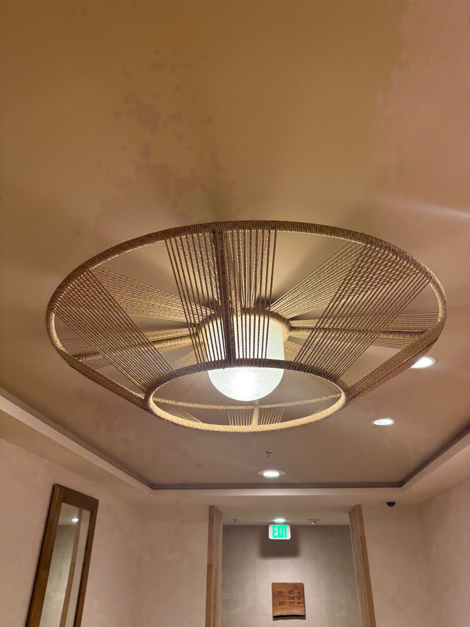 Ceiling light with woven shade in a hallway with beige walls and a green exit sign