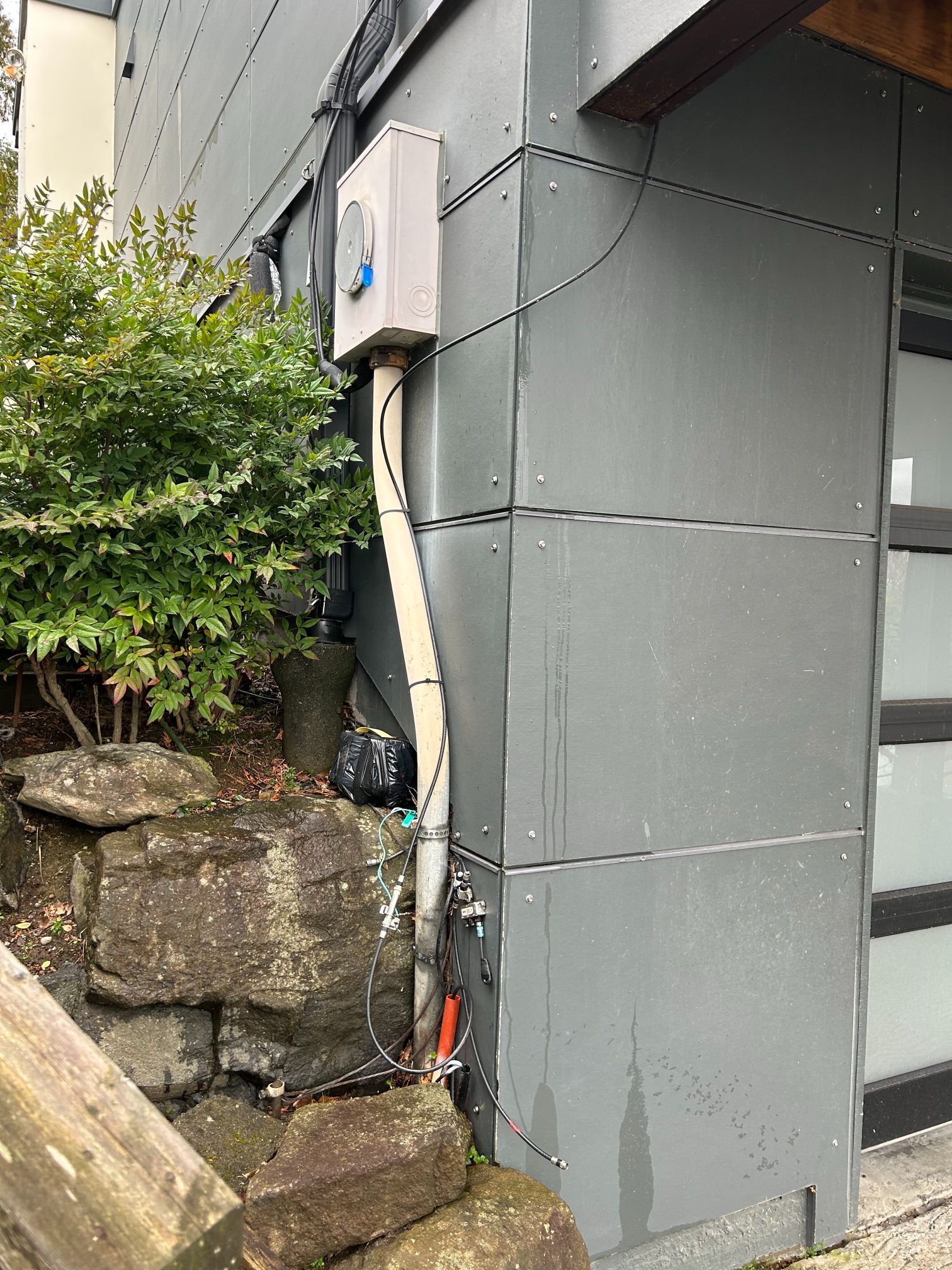Exterior wall with utility box, hanging cable, and stone steps beside greenery