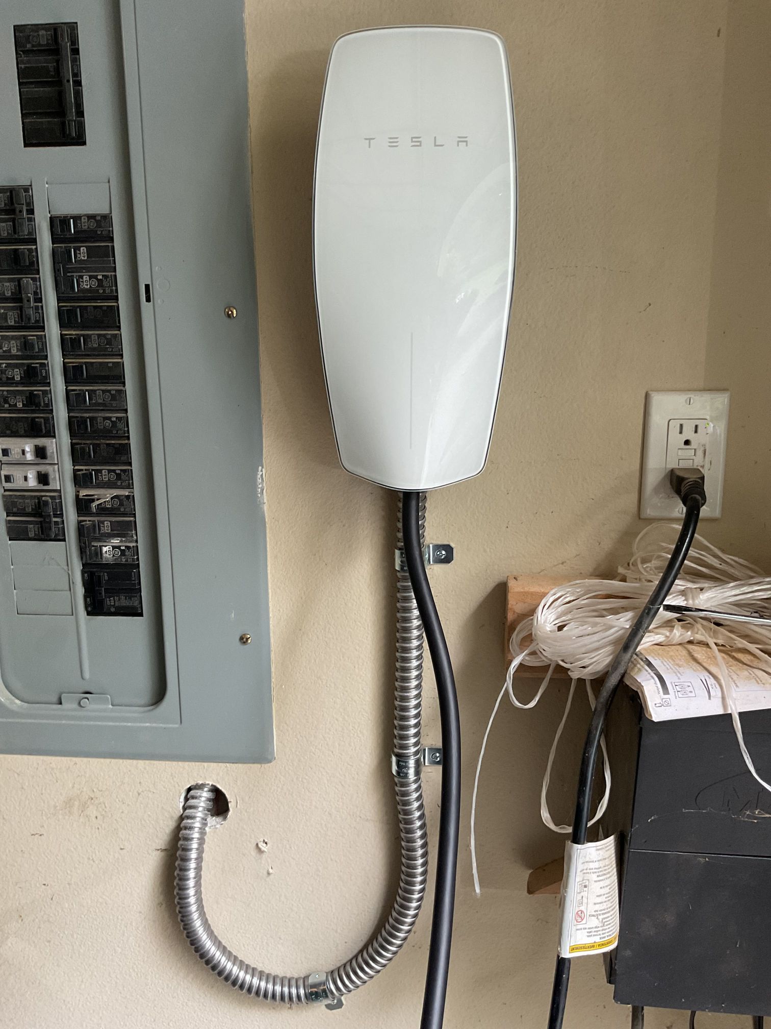 Wall-mounted white electric vehicle charger with coiled cable beside an electrical panel and outlet.