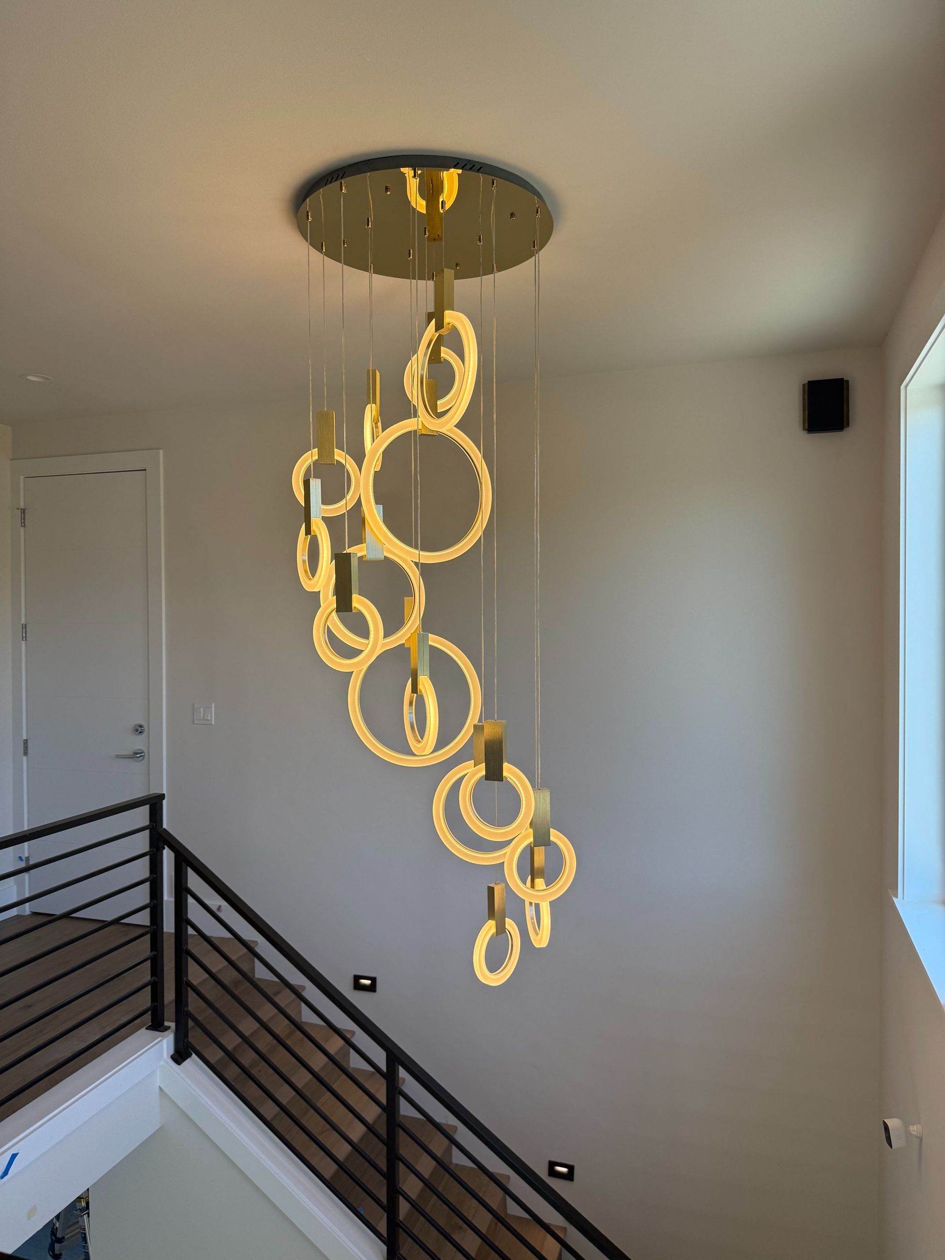 Modern gold ring chandelier hanging in a stairwell with black railings and white walls