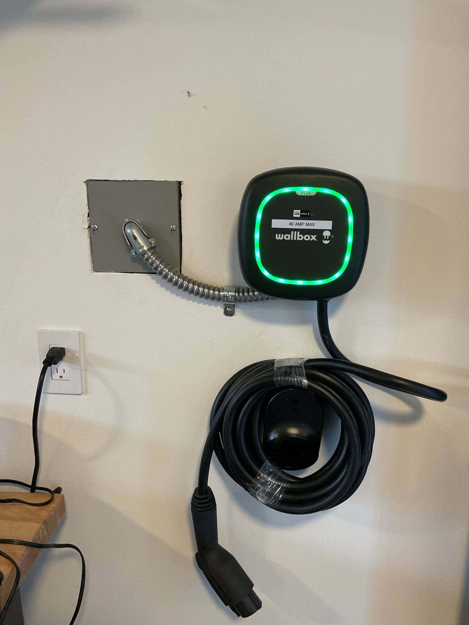 Wall-mounted EV charger with green light, coiled cable, and outlet on a white wall