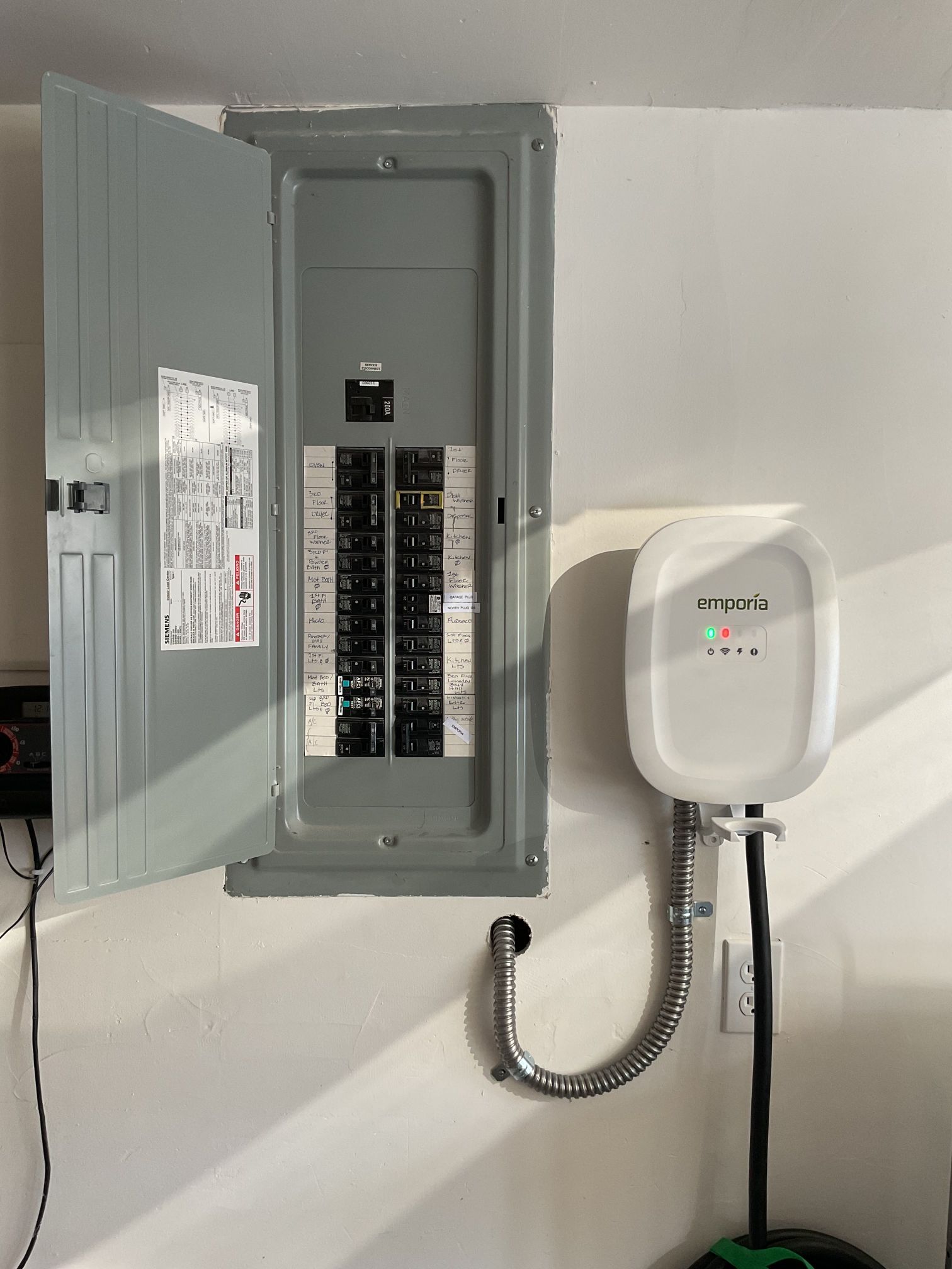 Open electrical panel with exposed breakers beside a white wall-mounted device and conduit