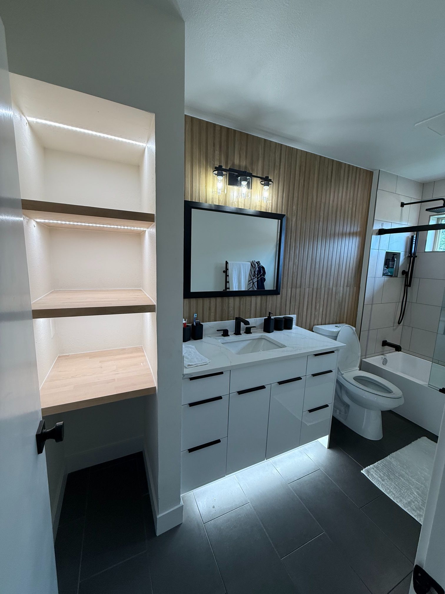 Modern bathroom with white vanity, mirror, toilet, walk-in shower, and built-in shelves with warm lighting