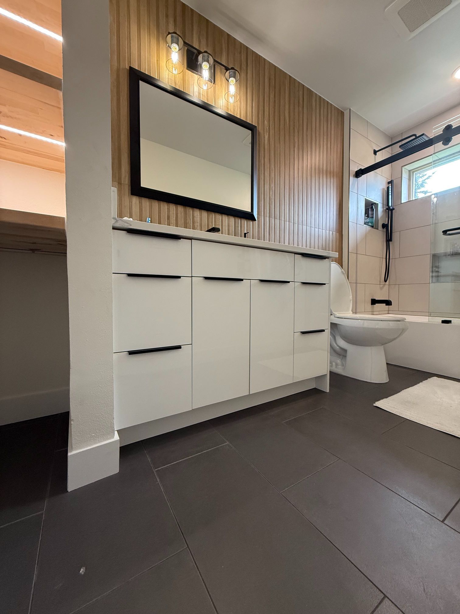 Modern bathroom vanity with white cabinets, wood wall paneling, mirror, sink, toilet, and dark tile floor.