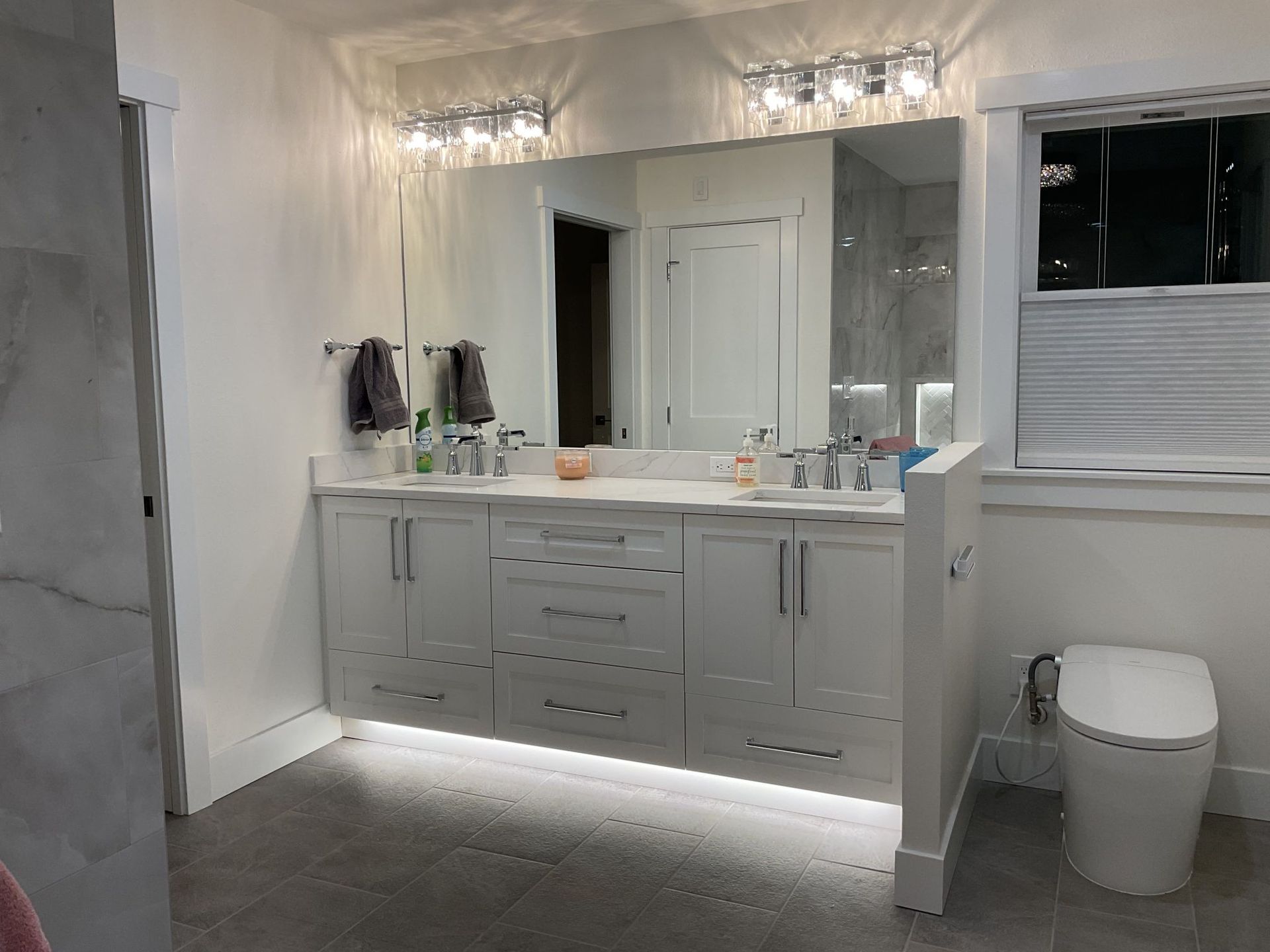 Modern white bathroom with double vanity, large mirrors, lit sconces, and a toilet in the corner