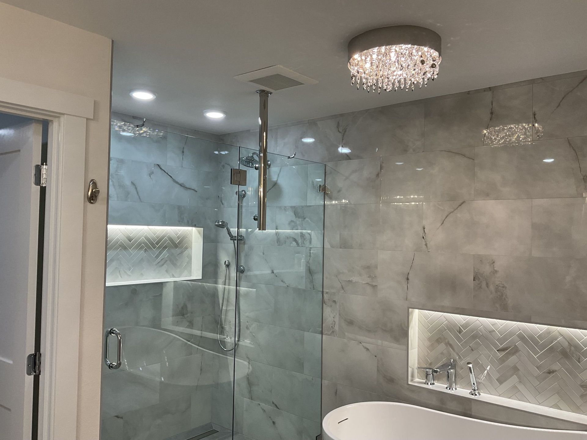 Modern bathroom with glass shower, gray tile walls, lit niches, and a crystal ceiling light