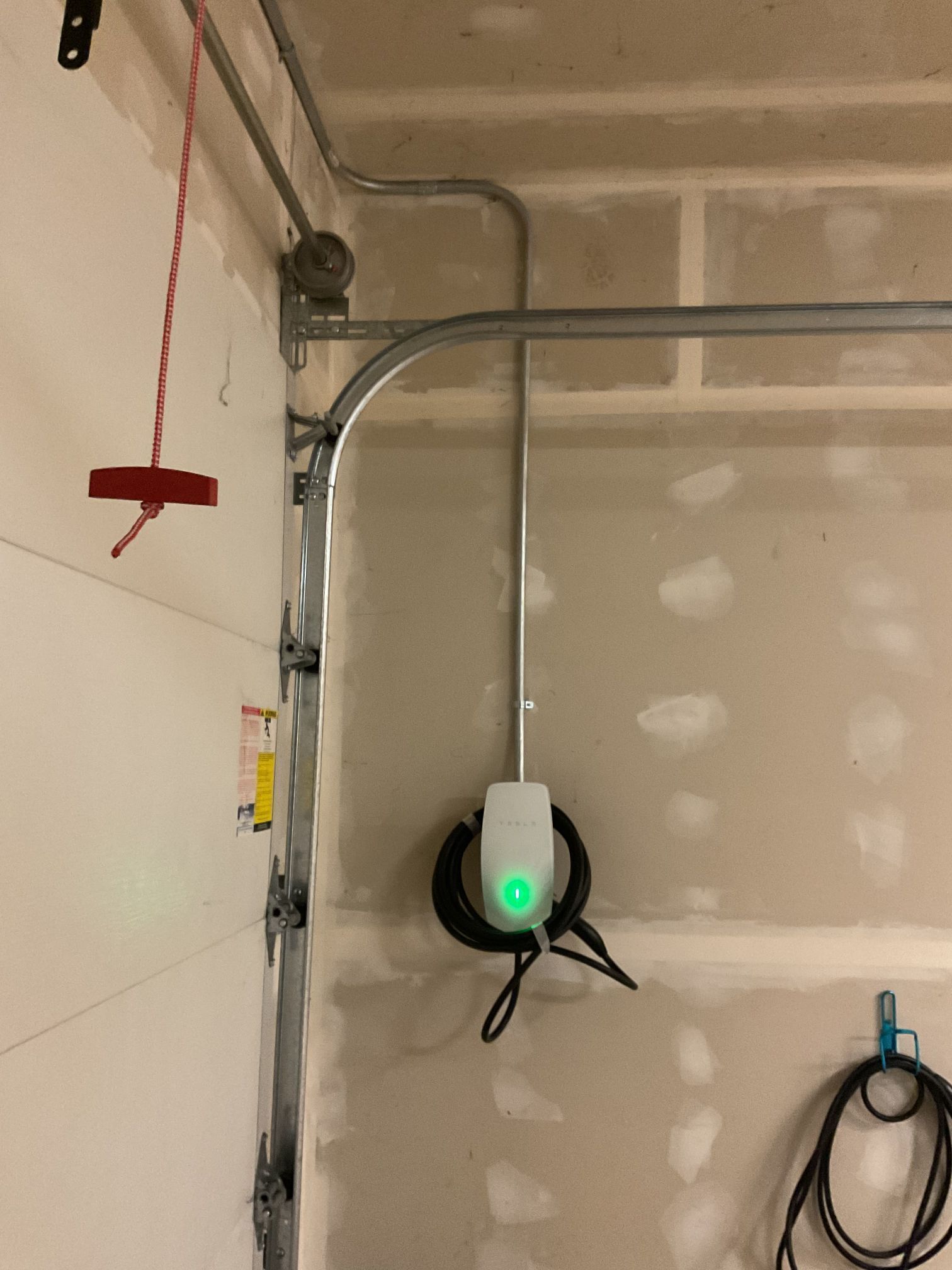 Wall-mounted EV charger with green light and coiled cable in a garage, with exposed conduit and utility items nearby