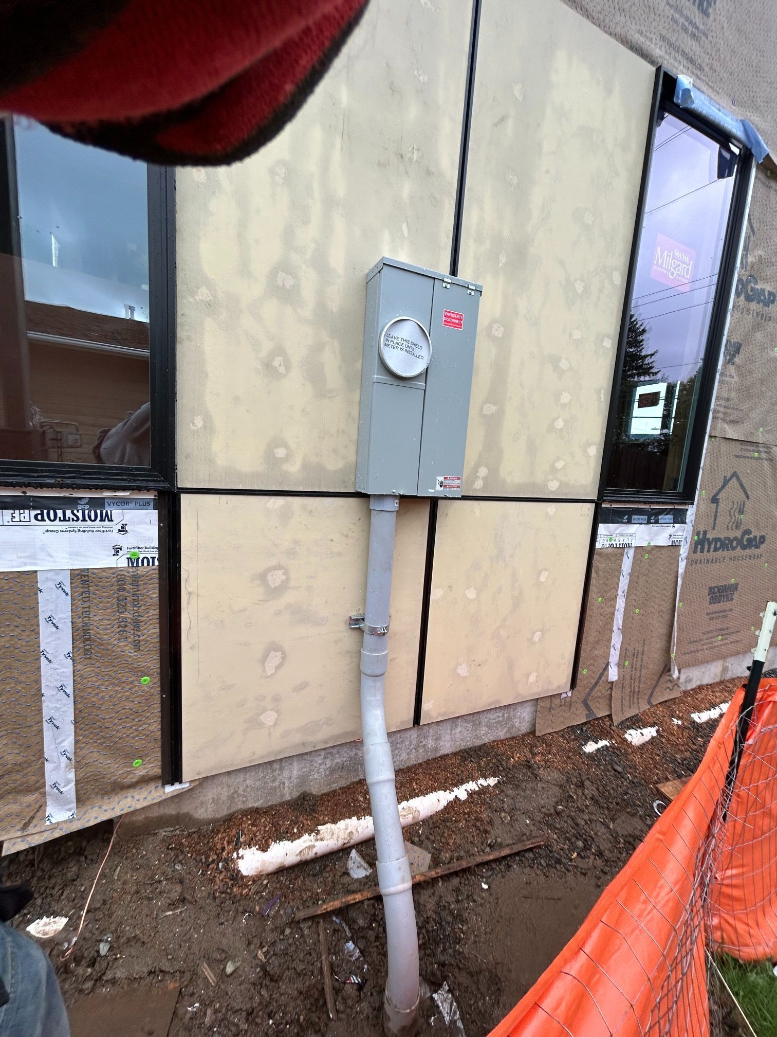 Gray electrical meter box on a building wall with conduit below, beside construction fencing and dirt ground
