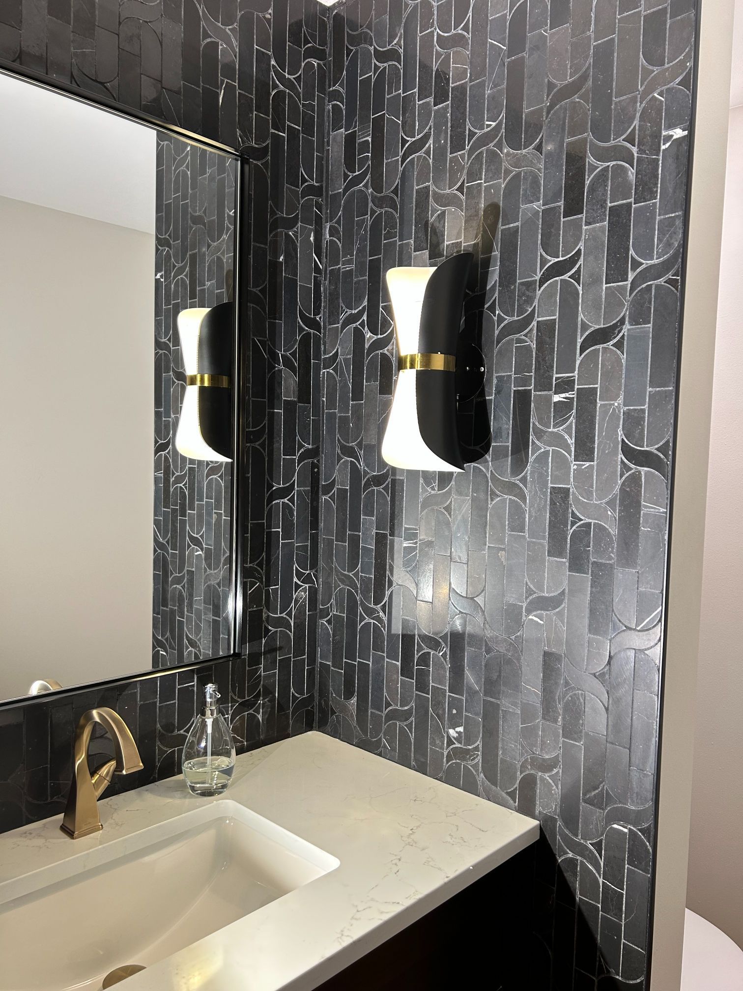 Modern bathroom vanity with white sink, gold faucet, and black geometric tile walls beside a mirror and sconces