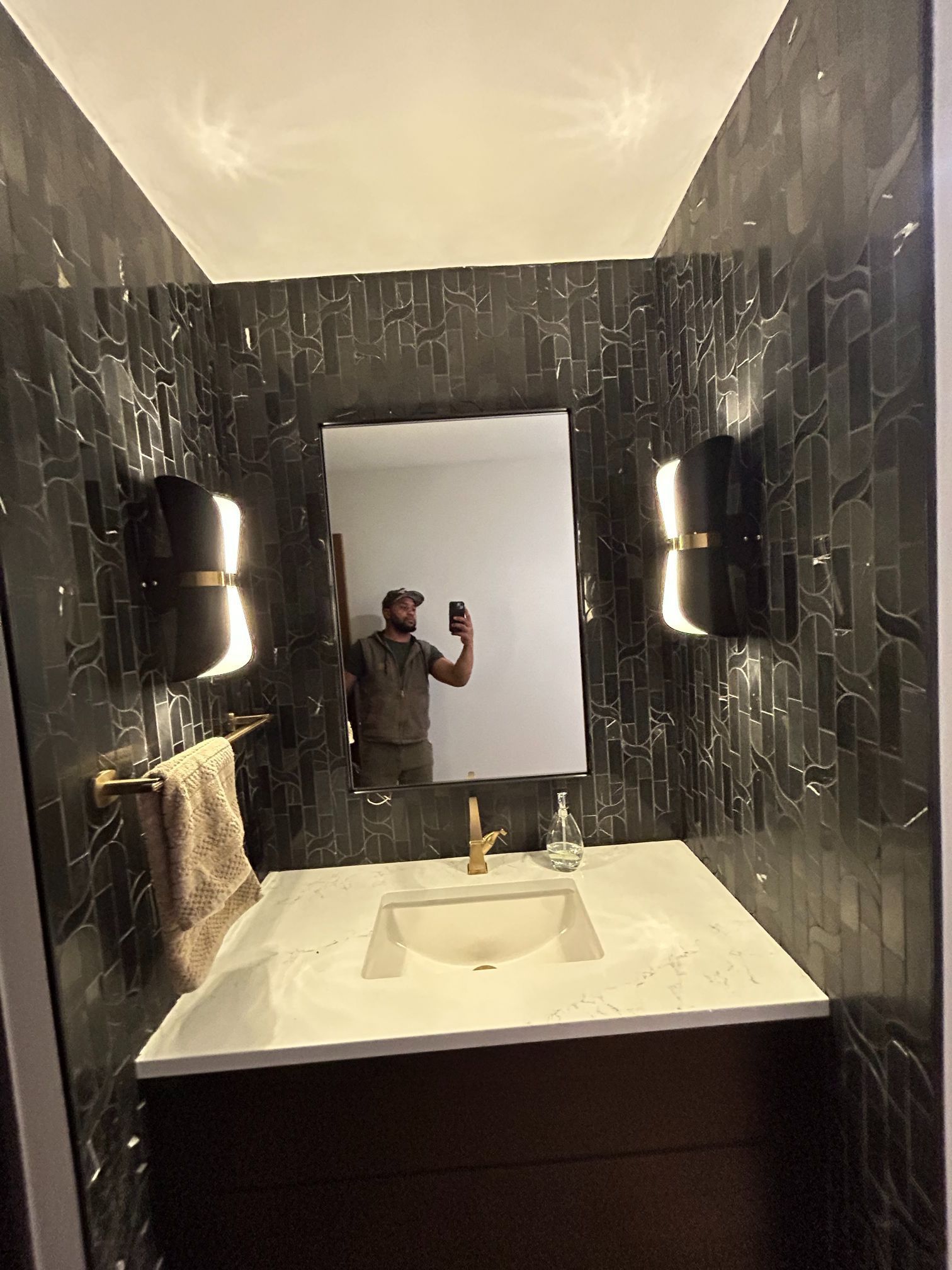 Dark-tiled bathroom mirror selfie with a white sink and vanity lights, showing a person holding up a phone.