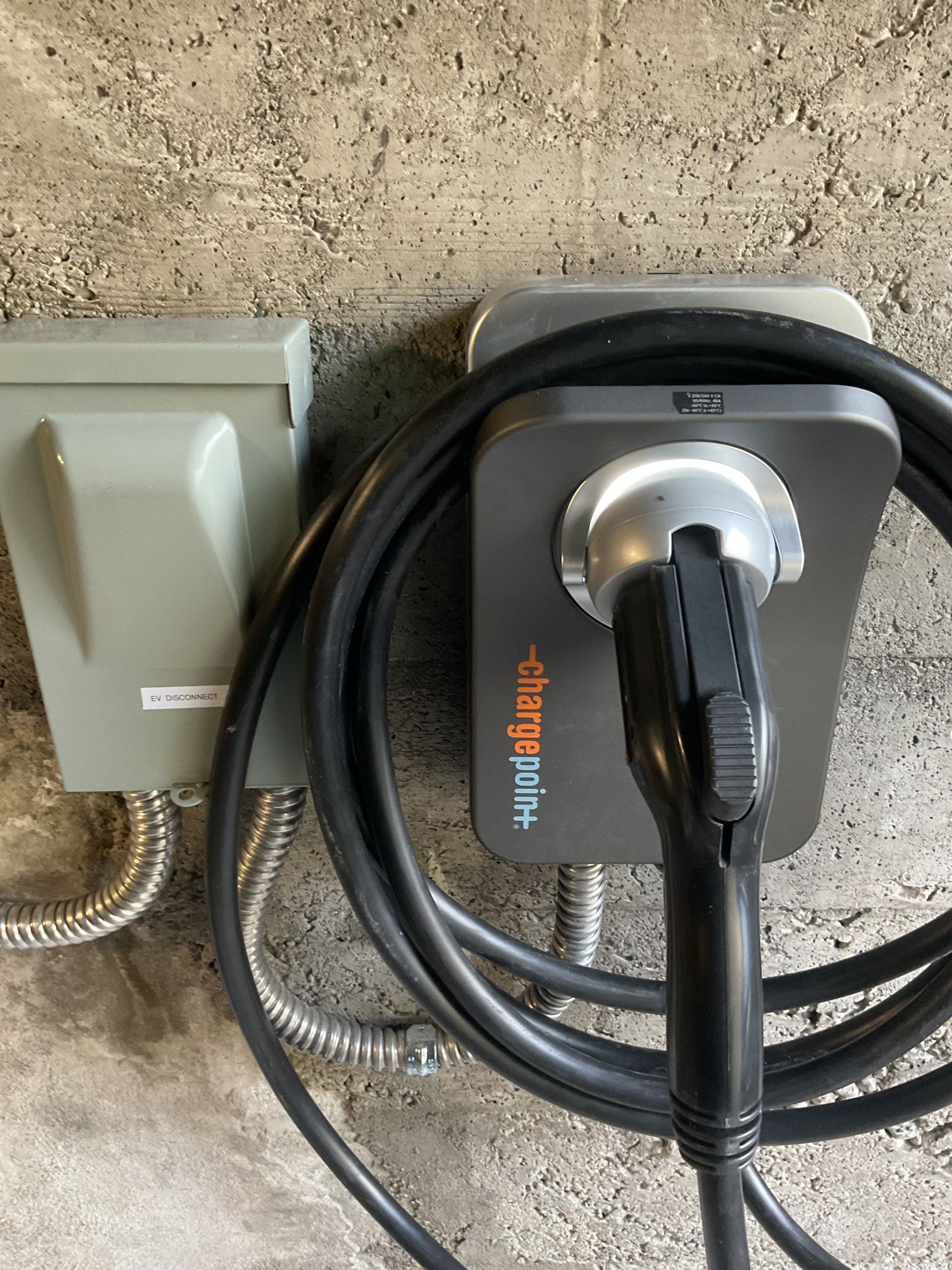 Coiled black charging cable plugged into a gray wall-mounted device next to an electrical outlet.
