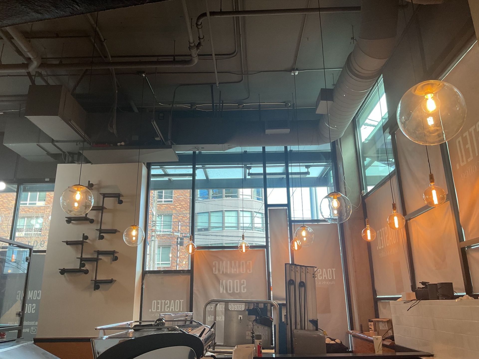 Modern café interior with hanging lights, large windows, exposed ceiling, and tables by brick walls