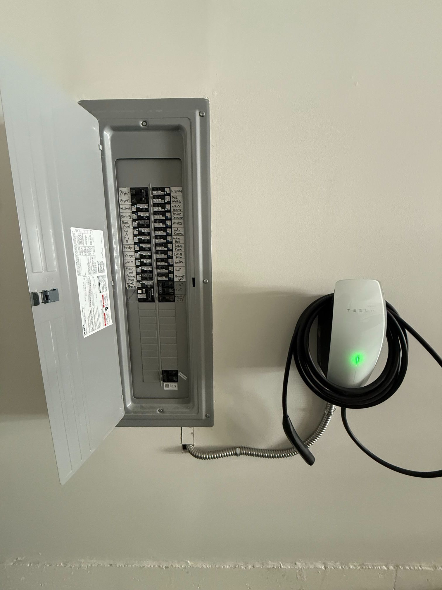 Electrical breaker panel with open door next to a wall-mounted charger with a green light and coiled cable.