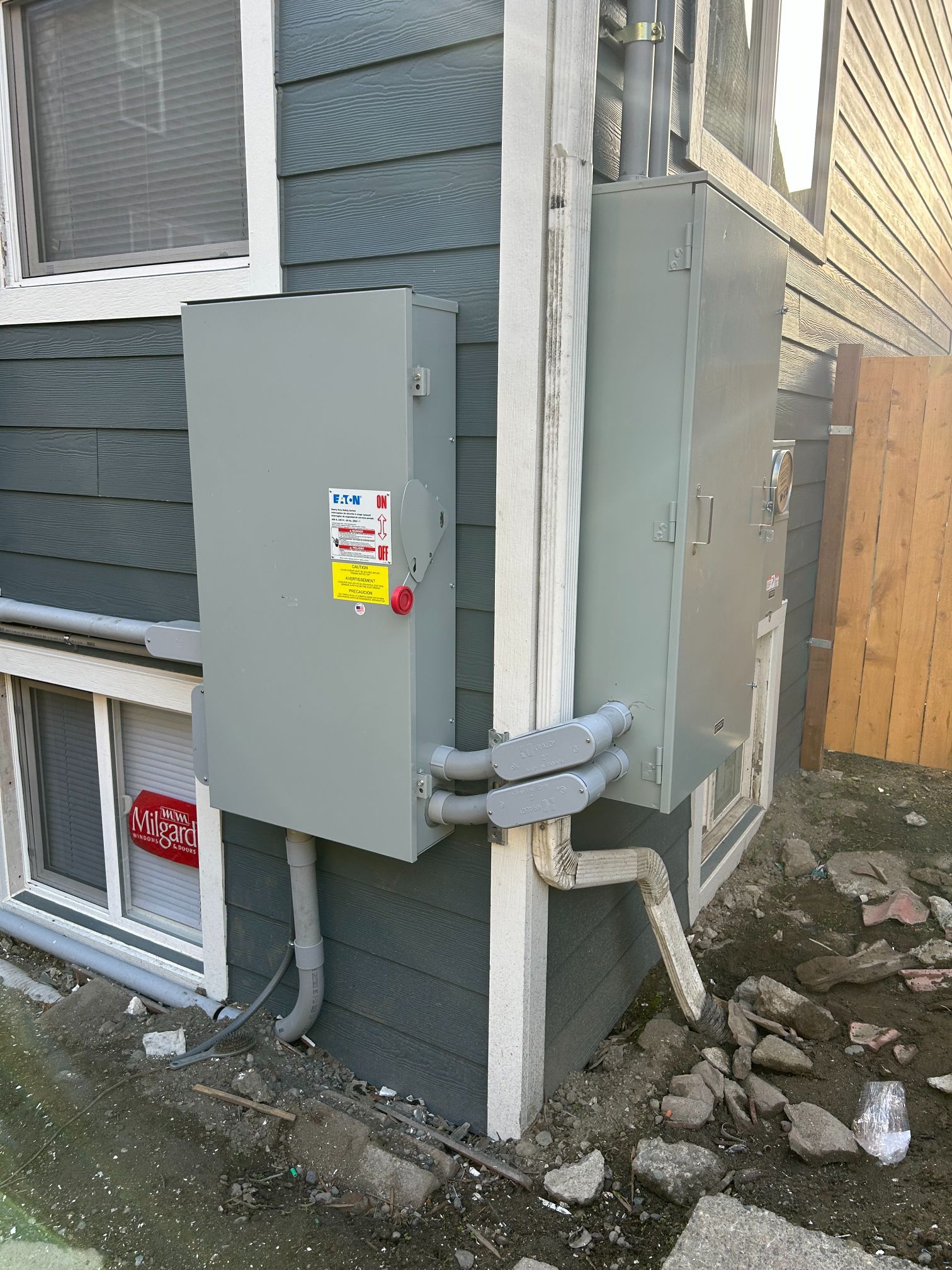 Gray electrical meter boxes mounted on a house exterior beside a window and utility pipes