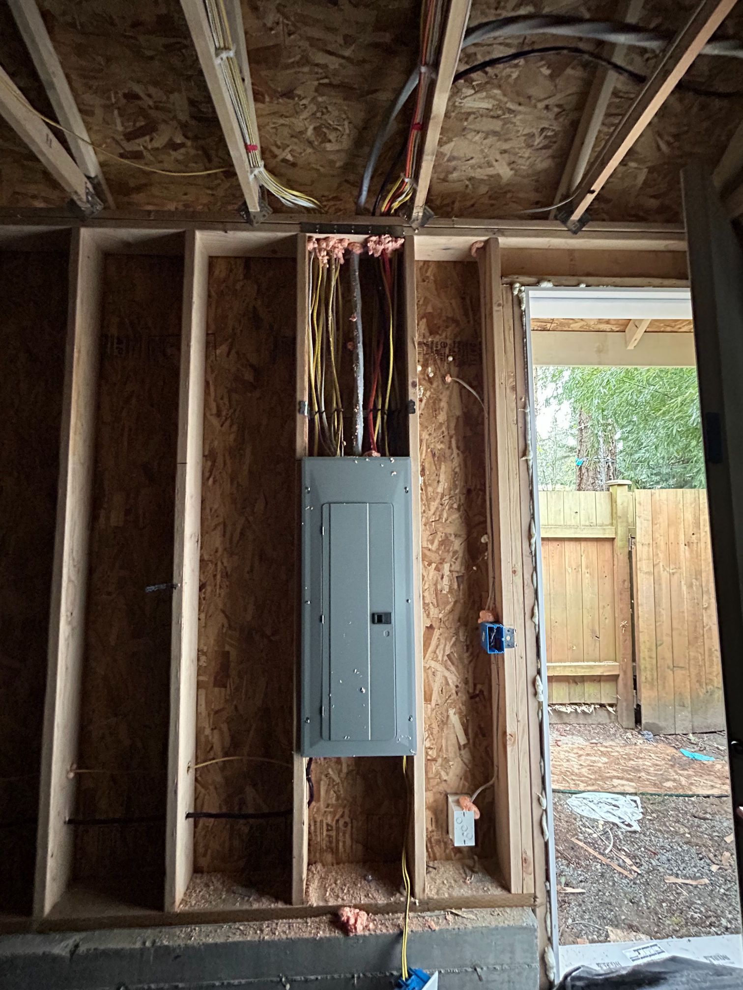 Exposed wall framing with an electrical panel and wiring in an unfinished room, doorway to a backyard visible.