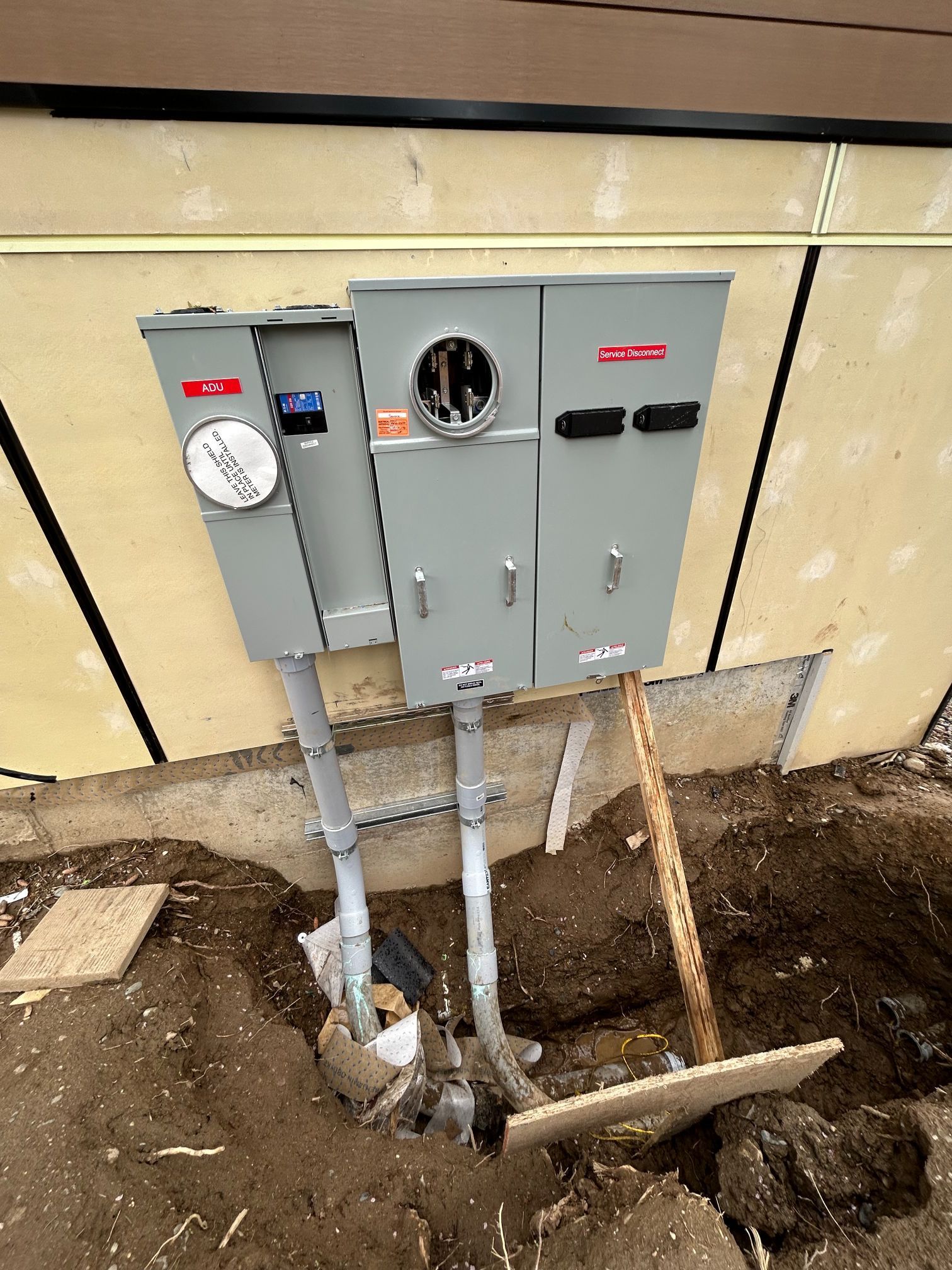 Electrical meter and breaker boxes mounted on an exterior wall above exposed trenching and conduit.