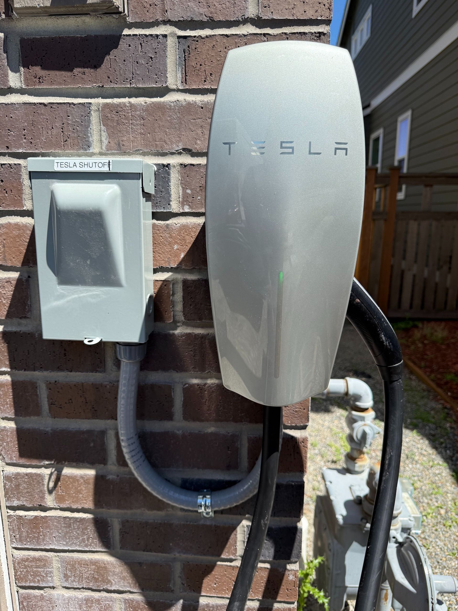 Tesla wall charger mounted on a brick exterior wall, with nearby electrical box and conduit.