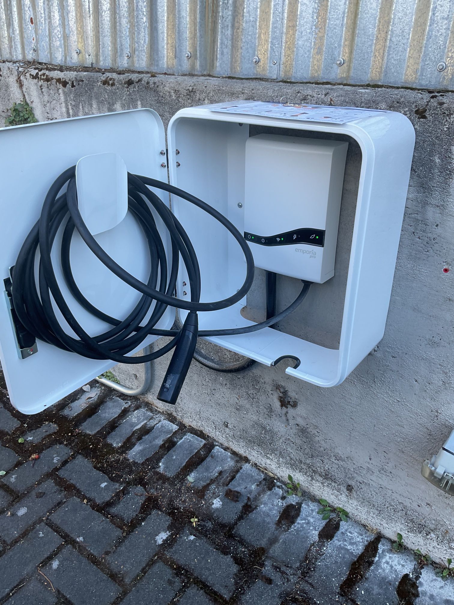 Open wall-mounted white electrical box with coiled black cables on a concrete walkway.
