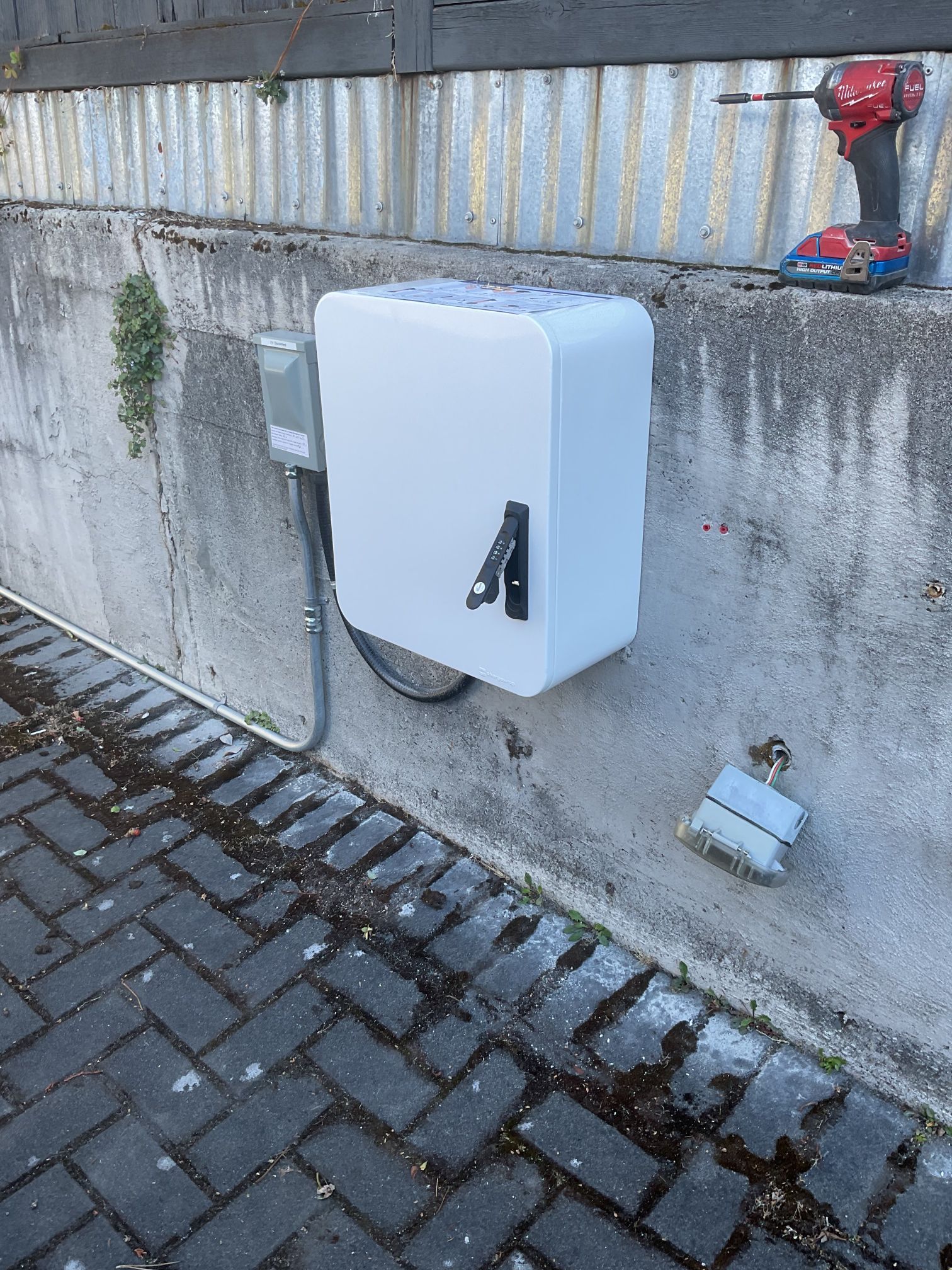 White utility box mounted on a concrete wall beside a paved walkway, with a red valve above it.