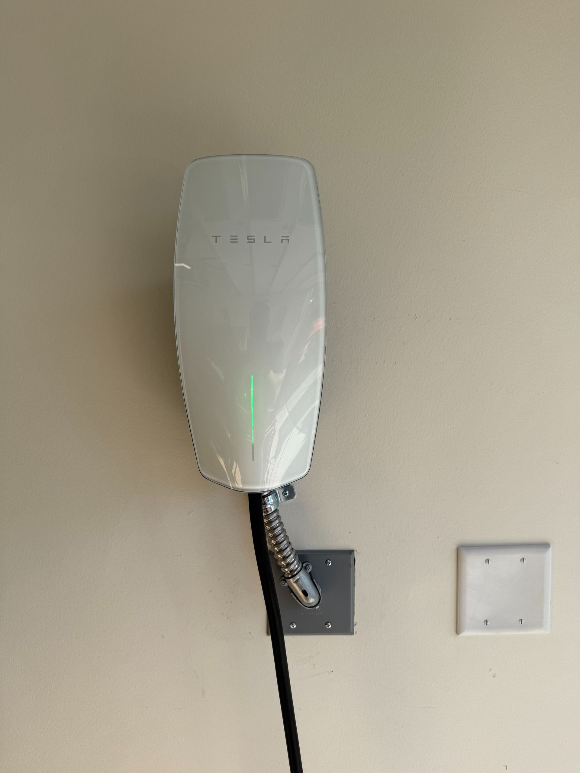 Wall-mounted white device with a green light and black cable on a beige wall, next to outlets.