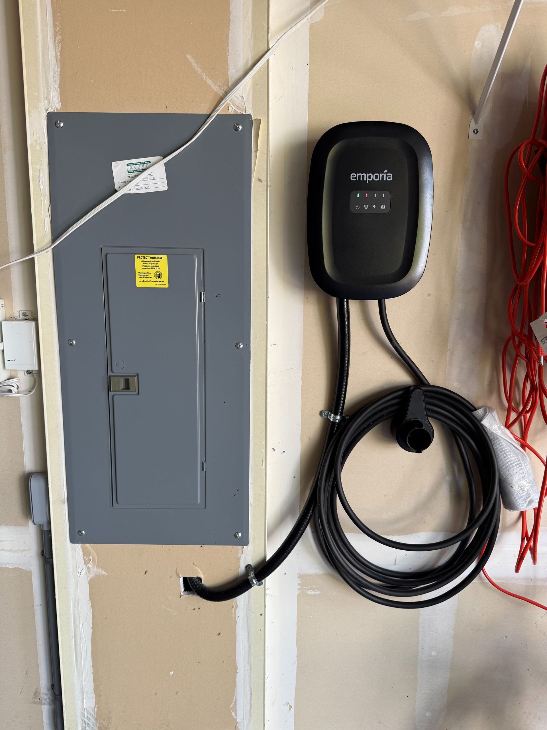 Gray electrical panel and black EV charger mounted on a beige wall with coiled cord and red cable nearby