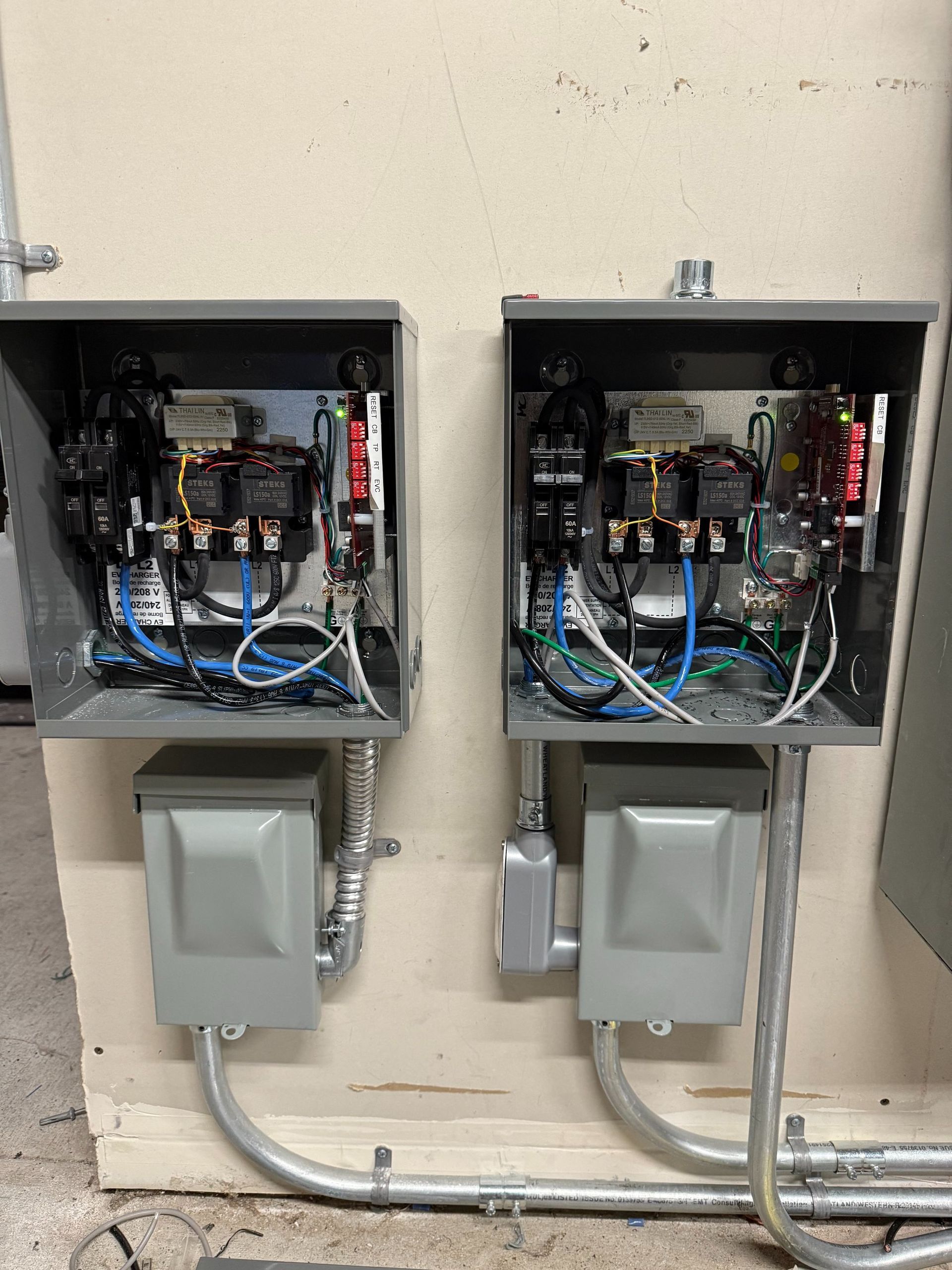Two open electrical breaker panels mounted above gray outdoor disconnect boxes on a wall.