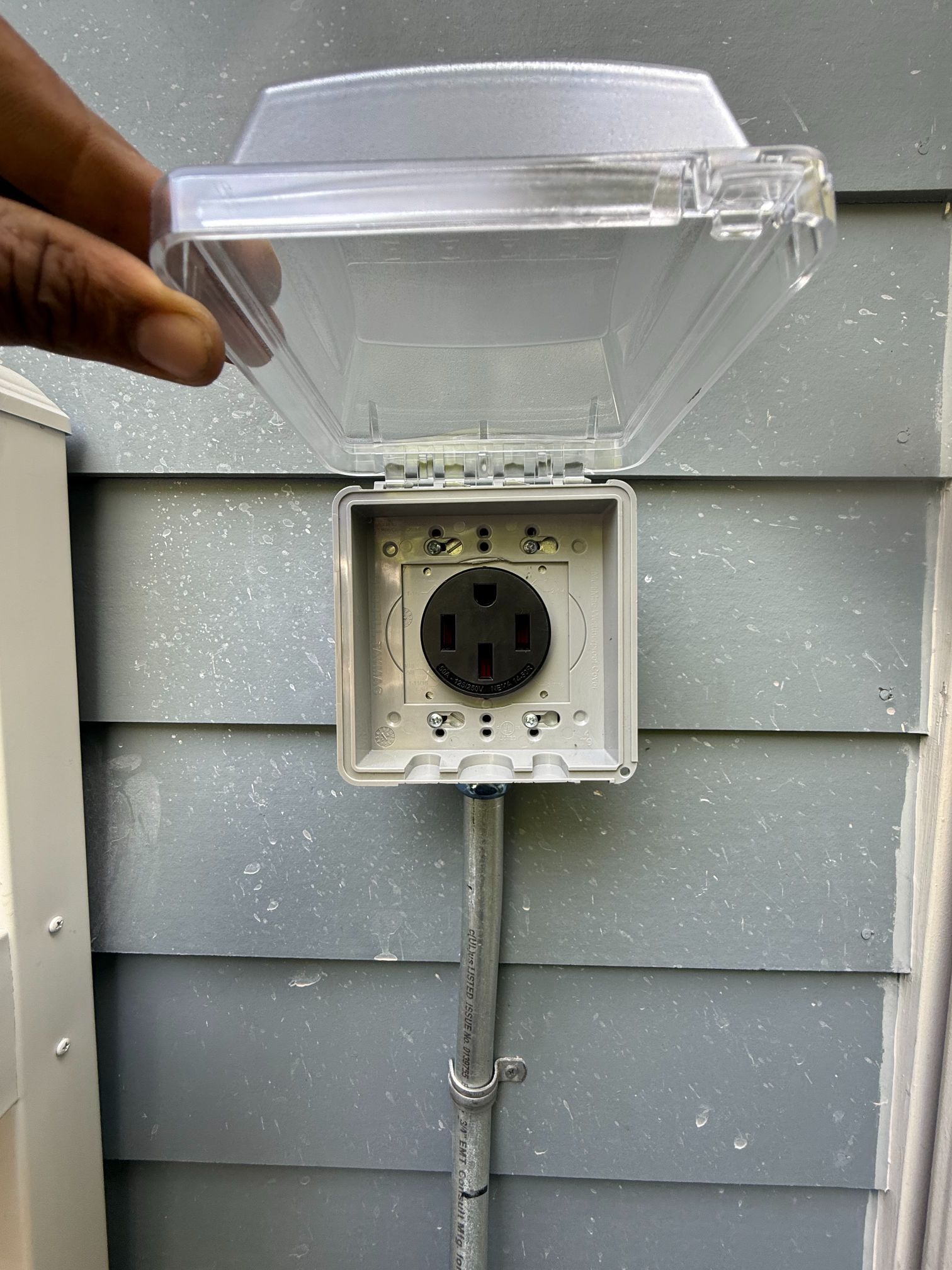 Hand holding a clear cover above an outdoor electrical outlet mounted on gray siding.