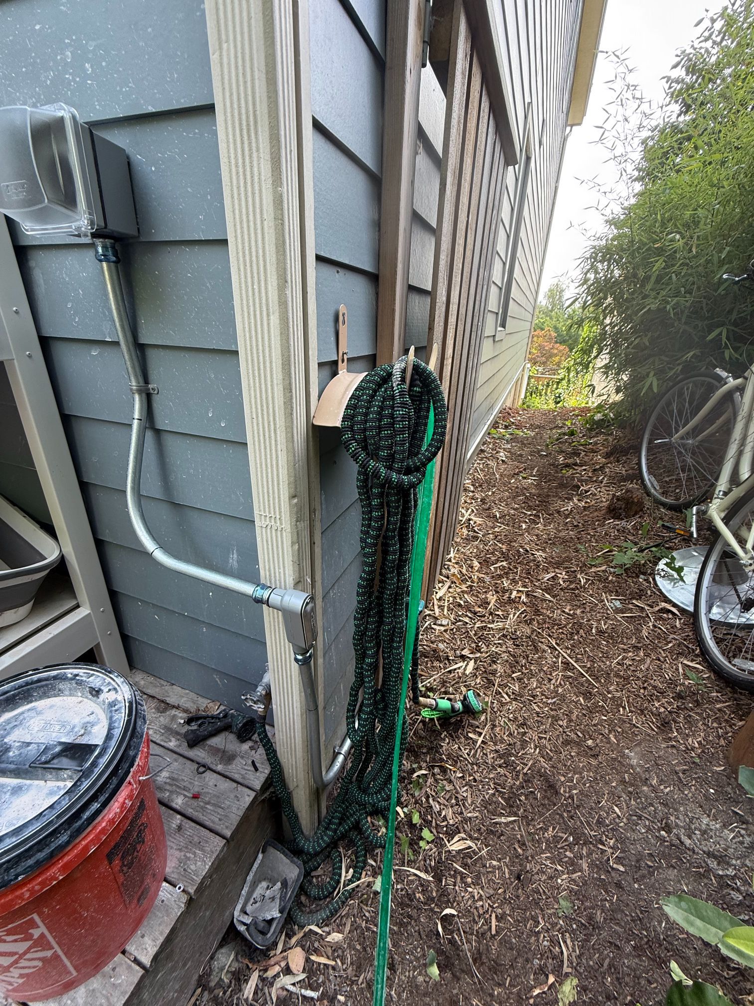 Green rope tied to a black post beside a house, with a red bucket and narrow dirt path nearby.