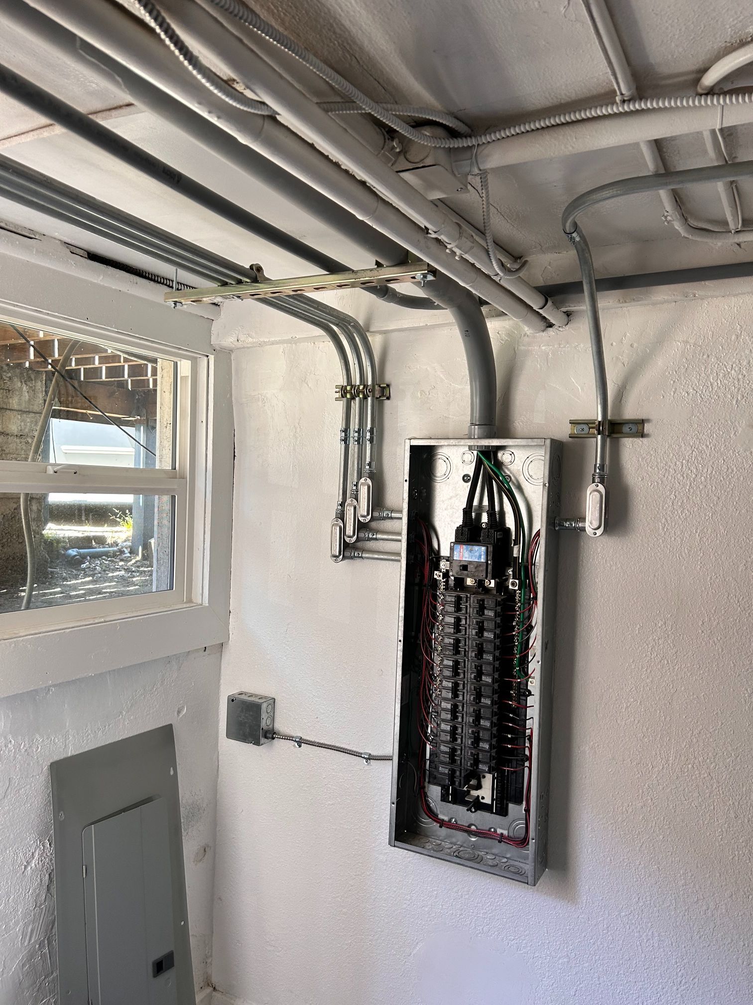 Electrical panel and exposed conduit in a utility room with a window and open ceiling.