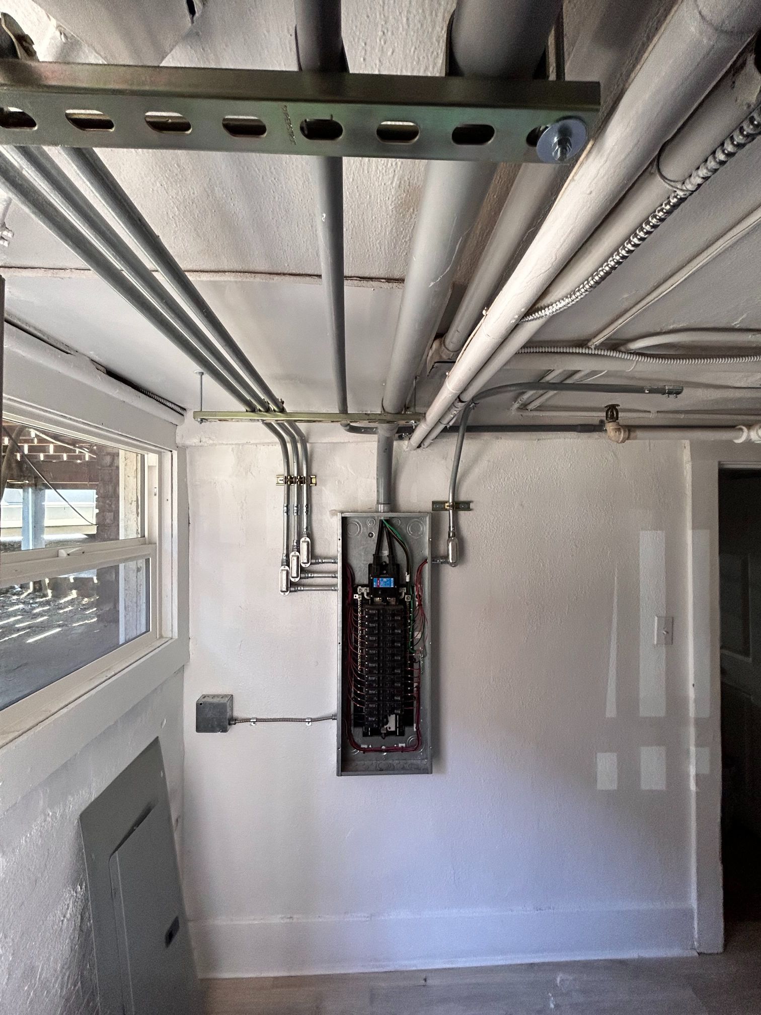 Electrical panel in a utility room with exposed ceiling pipes and metal framing.