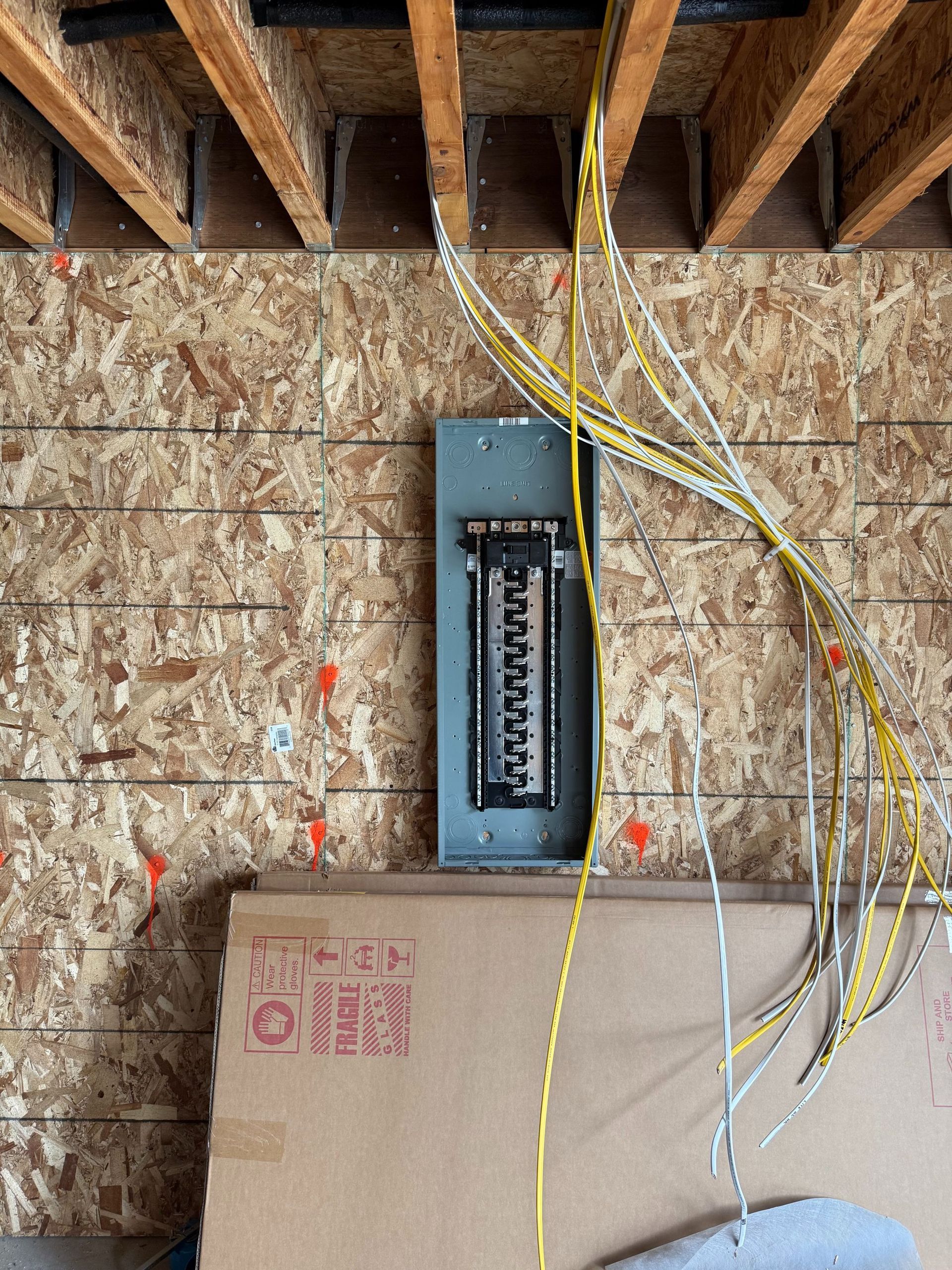 Open electrical panel mounted on plywood wall under exposed joists, with yellow and white wires hanging nearby