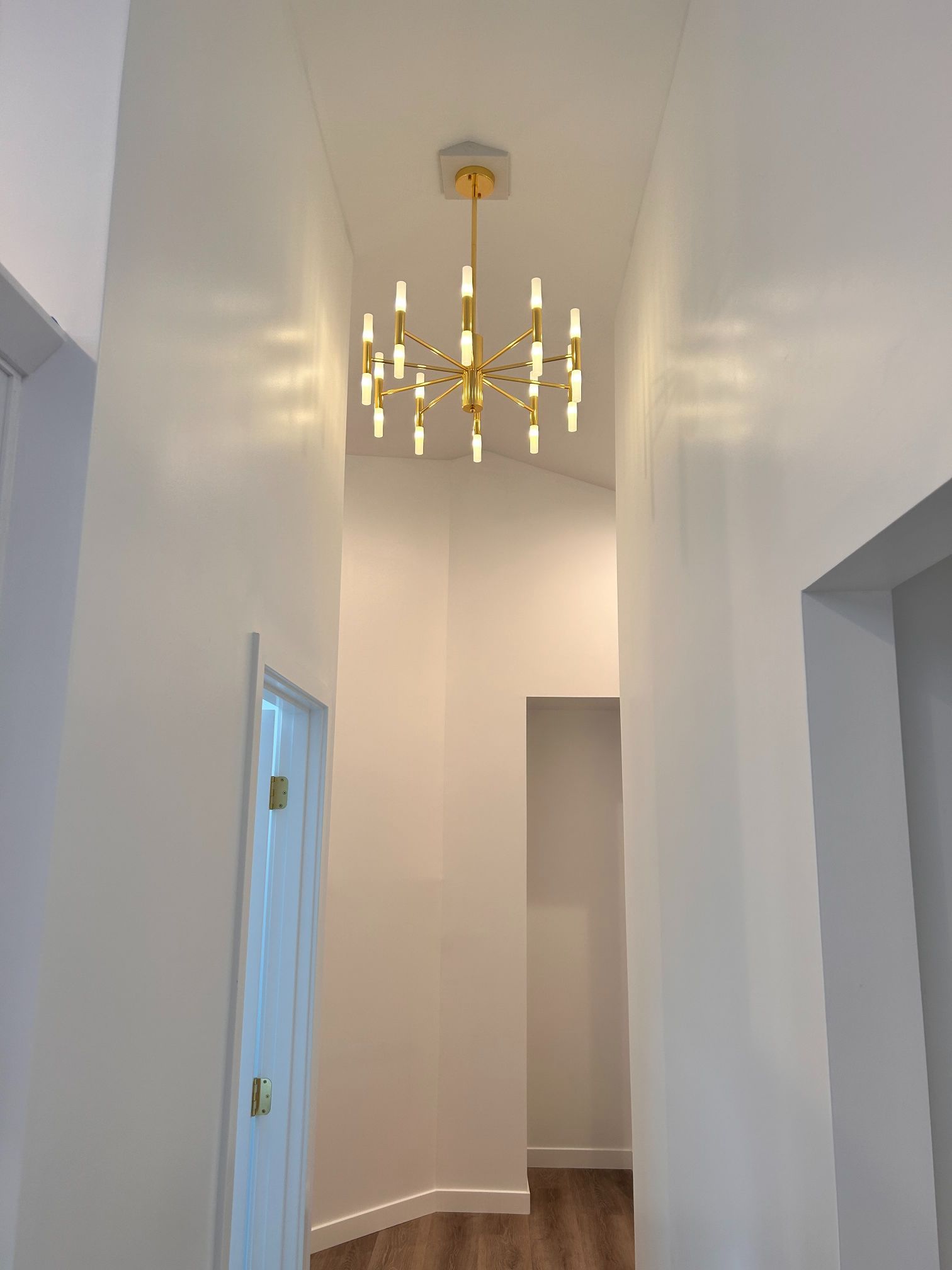 Narrow hallway with white walls, wood floor, and a gold chandelier hanging from the ceiling
