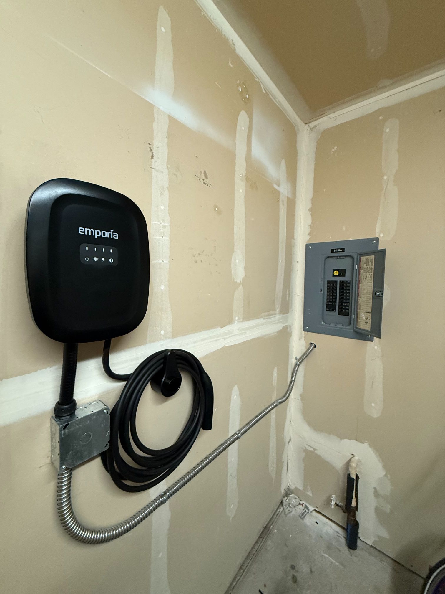 Black EV charger mounted on drywall next to an electrical panel in a garage-like room.