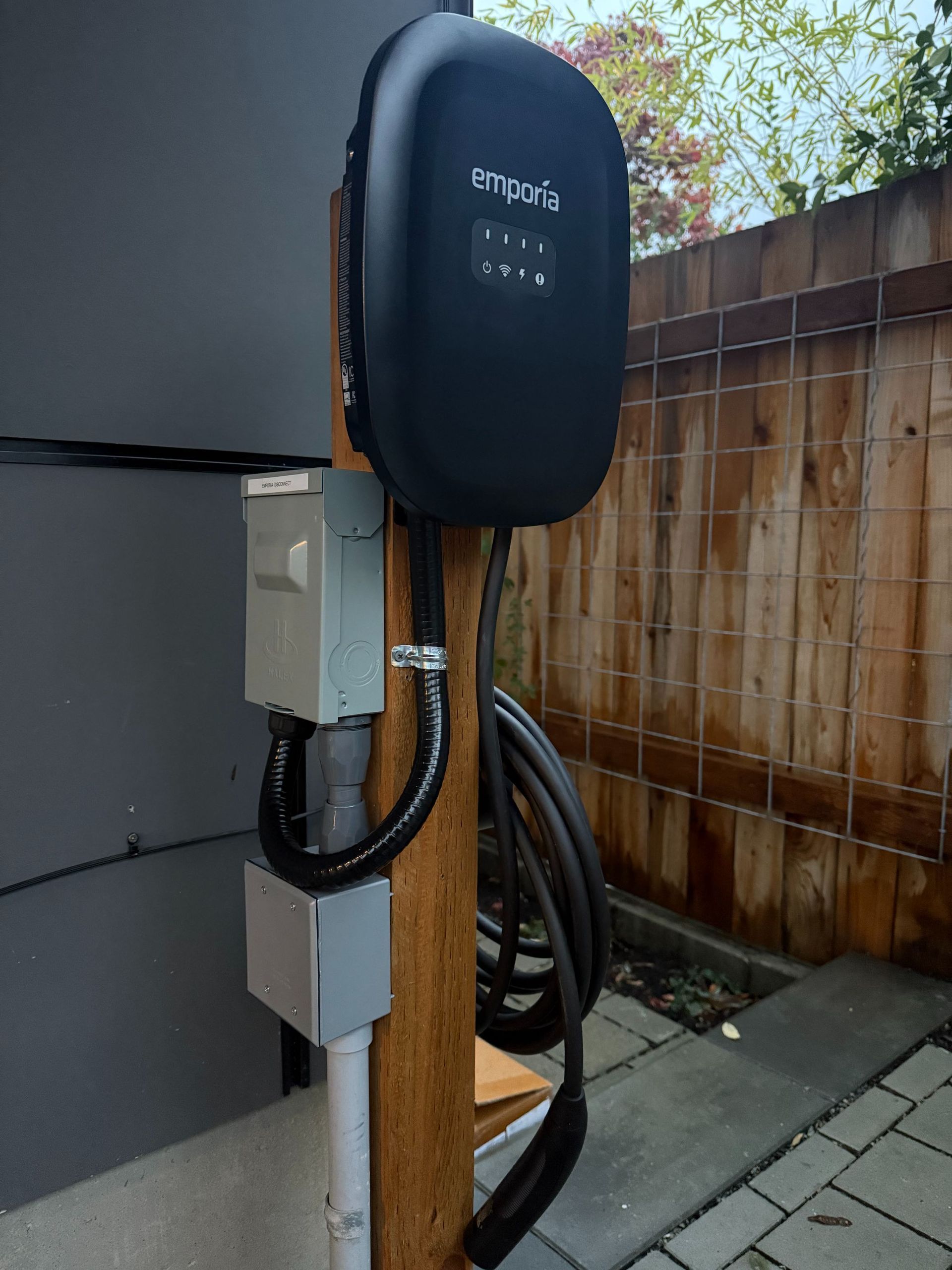 EV charger mounted on a wooden post beside a house and fence, with cables coiled below.
