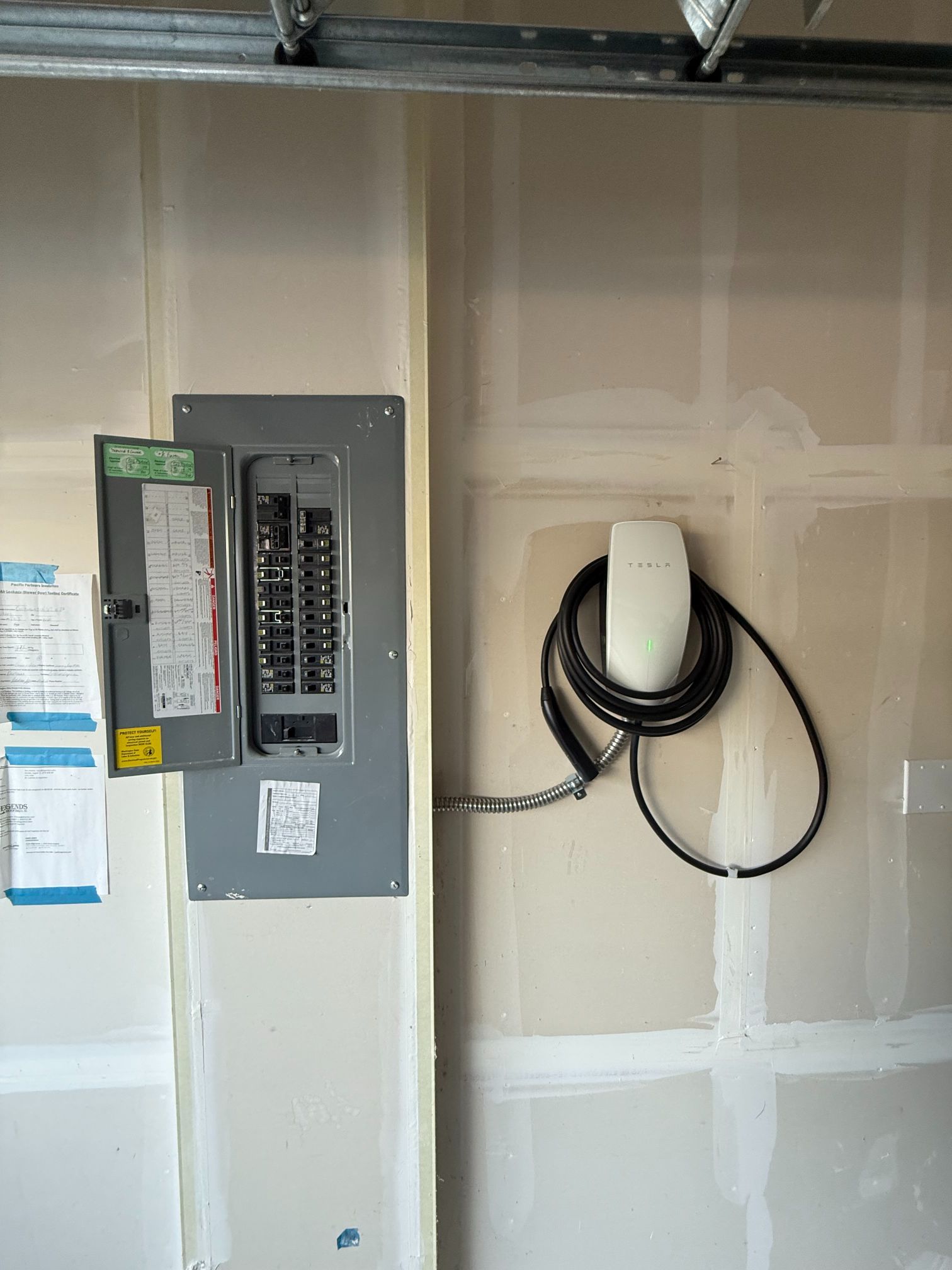 Electrical panel and coiled black cable mounted on unfinished drywall wall.