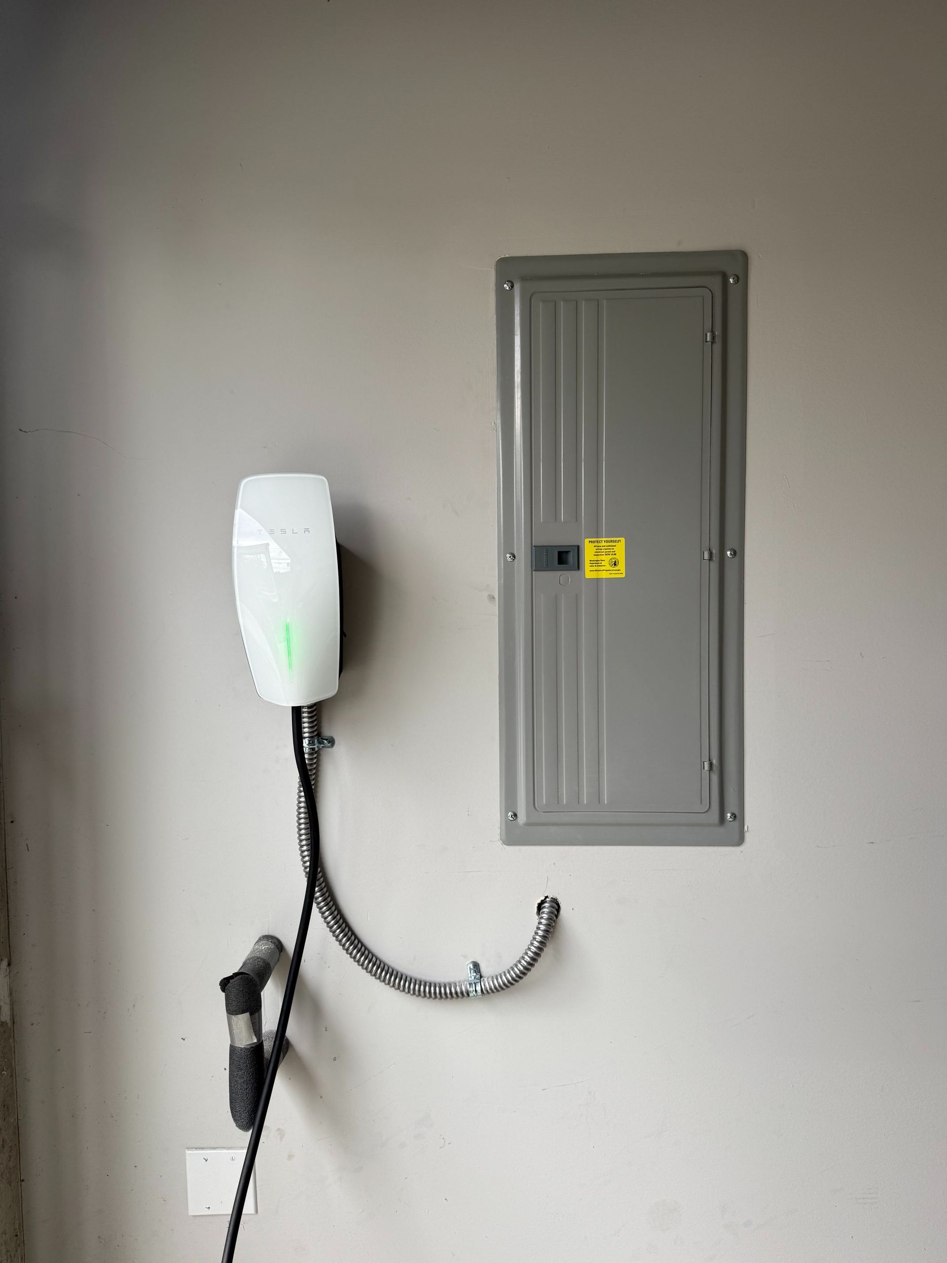 Wall-mounted charging station with a white device and black cable beside a gray electrical panel.