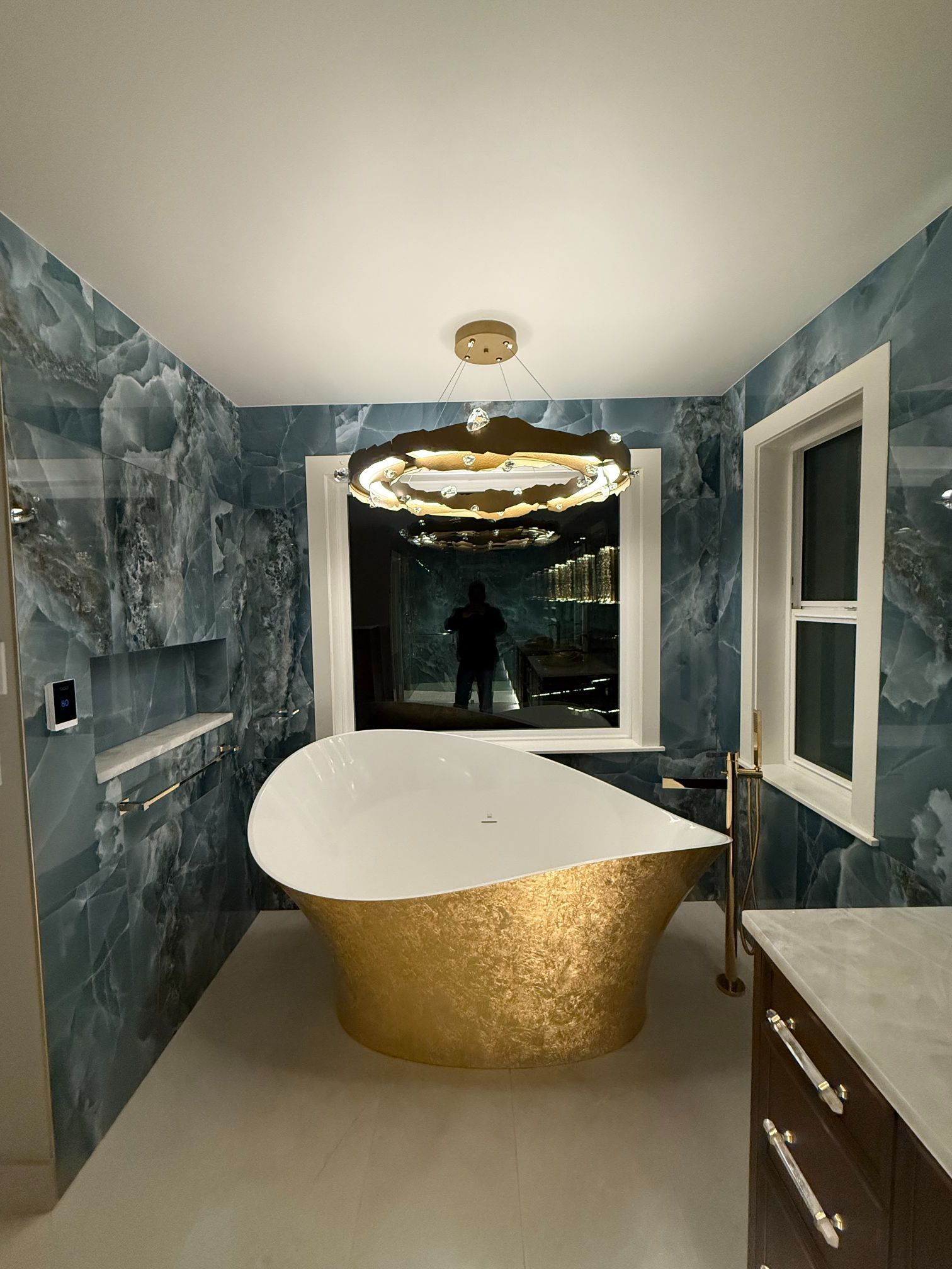 Modern bathroom with blue marble walls, gold tub, large round mirror, and window view of city lights