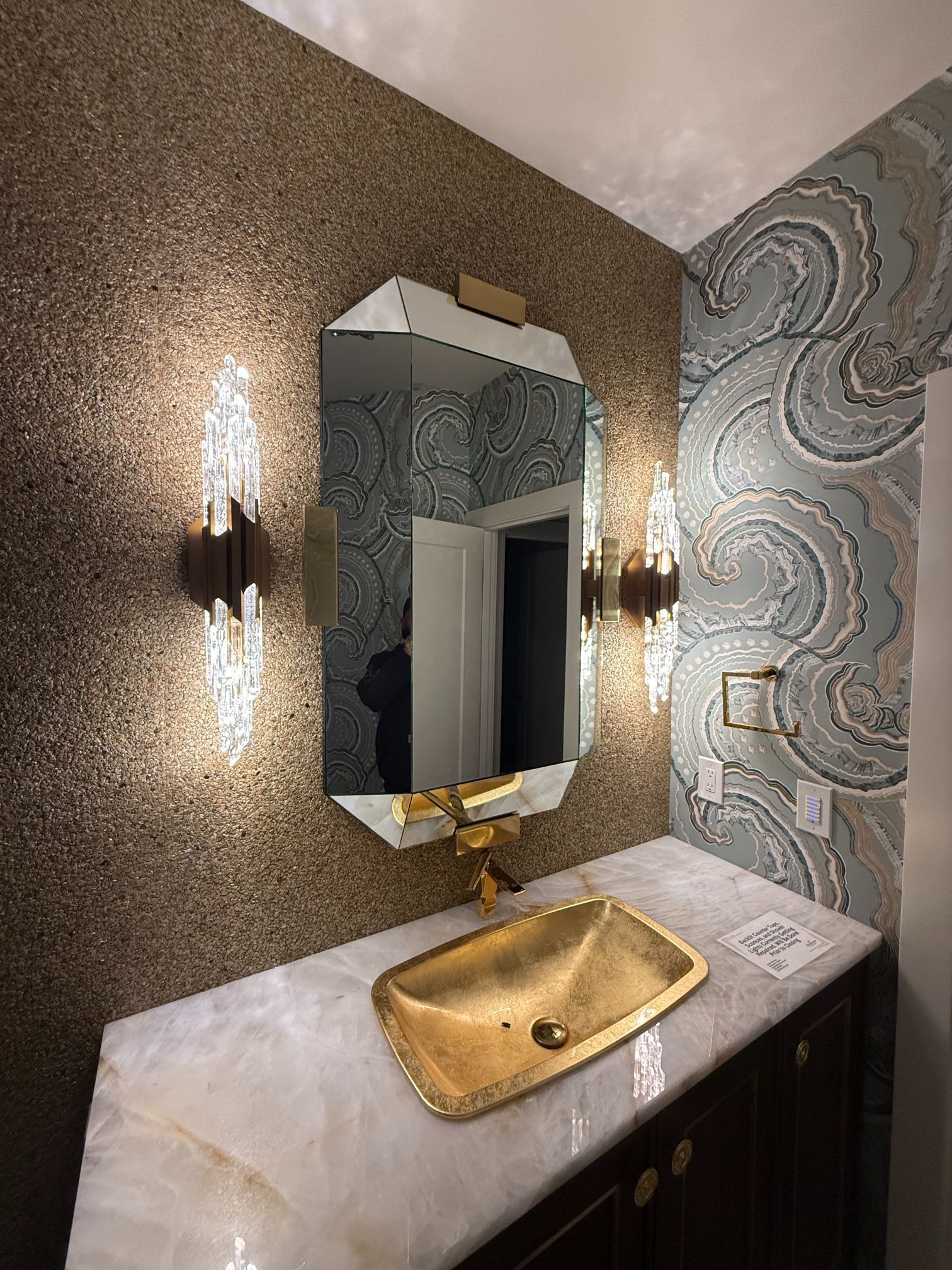 Gold sink and mirror in a small ornate bathroom with patterned blue wall tiles and sconces