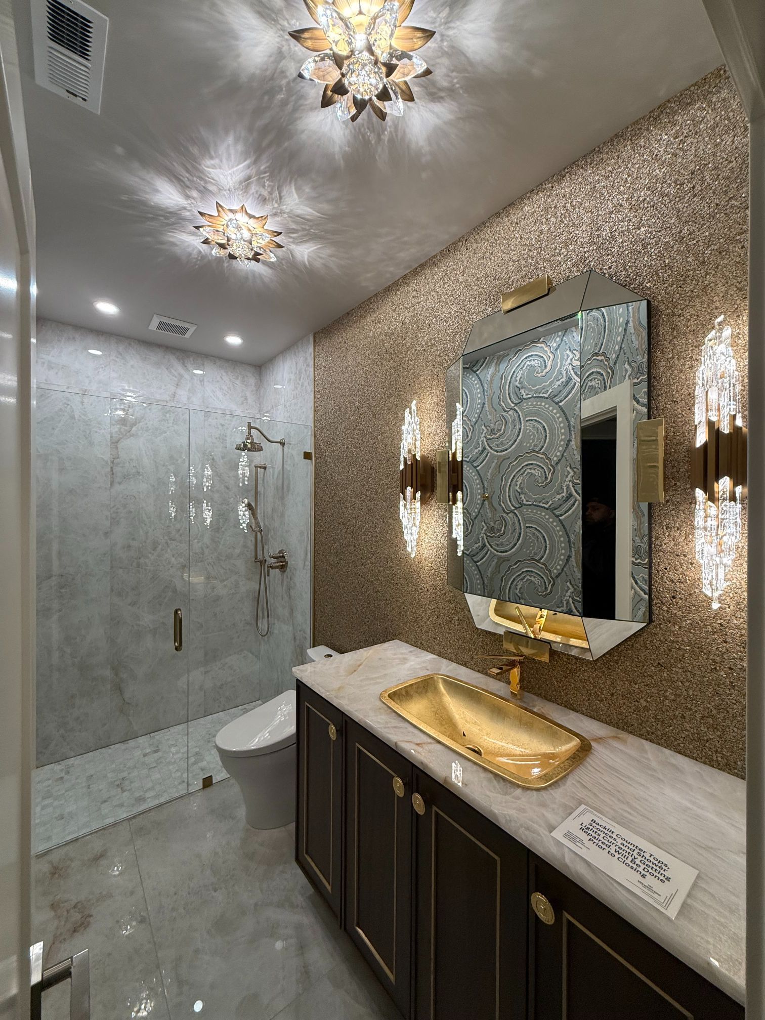 Modern bathroom with glass shower, mosaic walls, dark vanity, gold sink, and ornate ceiling lights